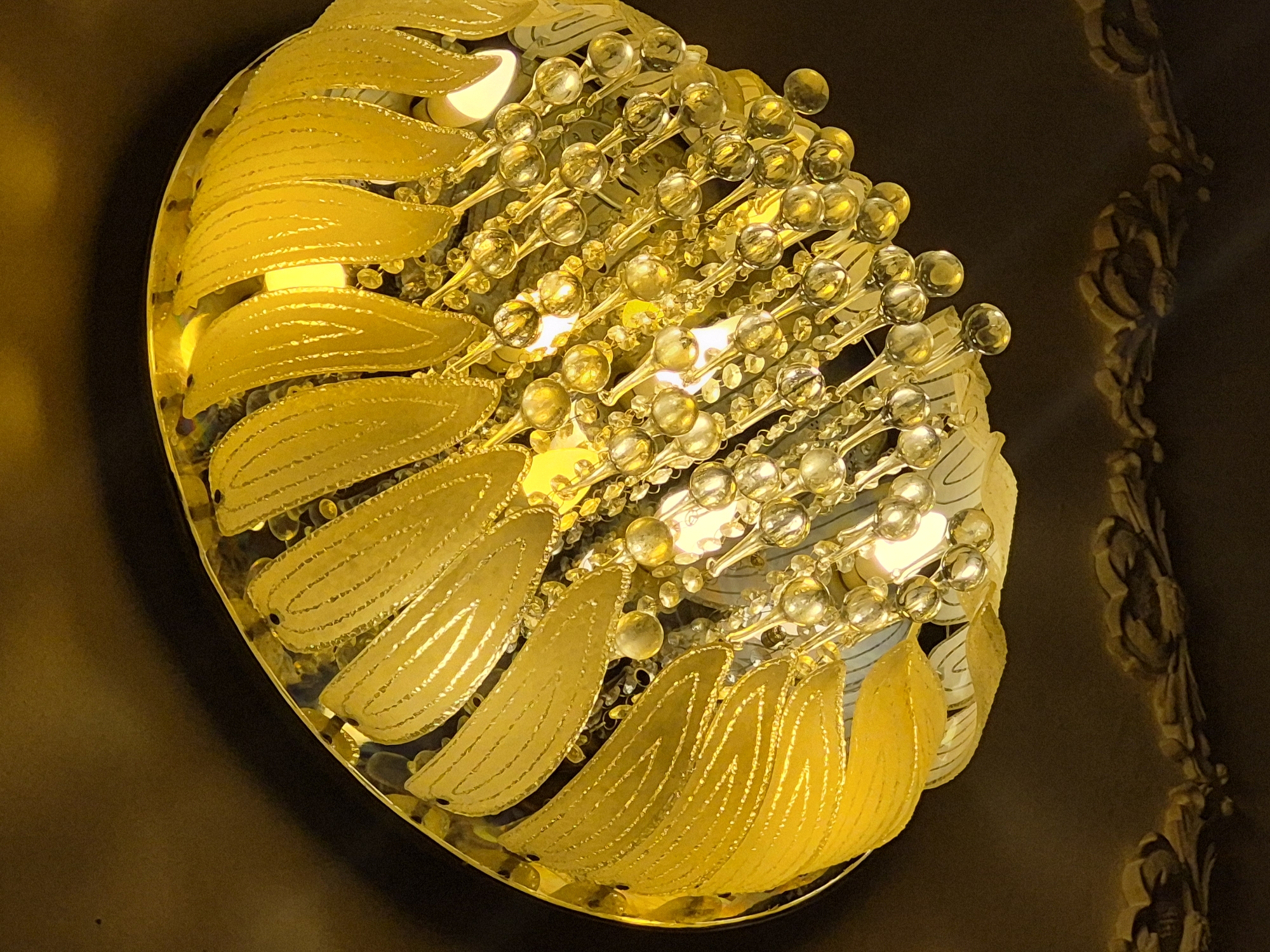 The image displays an opulent, illuminated light fixture, likely a chandelier or ceiling lamp, seen from a slightly angled, overhead perspective. The fixture is dominated by warm golden and yellow hues, radiating a bright glow against a dark background.
The central part of the fixture features numerous small, clear and golden, spherical or teardrop-shaped glass beads or crystals. These are clustered together on delicate stems, reflecting and refracting the intense yellow light emanating from internal sources. Surrounding this glittering core is a layered arrangement of translucent, leaf-like or petal-shaped elements. These larger pieces are also golden-yellow and exhibit a subtle textured pattern of fine, parallel lines, suggesting intricate craftsmanship. They appear to be made of glass or a similar material, allowing light to pass through them and illuminate their form.
Several bright, warm white and yellow light bulbs are visible within the fixture, providing the primary illumination. The background is a deep, dark brown or black, which emphasizes the brilliant glow of the light fixture. On the far right edge of the image, a faint, blurred, and shadowy pattern or reflection of an ornate, possibly carved or molded vertical structure is visible on the dark surface, hinting at detailed surrounding decor or another part of the fixture.
There are no people or specific activities depicted. The setting is indoors, with the illuminated fixture suggesting it is evening or night. No text is visible in the image. The overall impression is one of elegance, luxury, and intricate design.
