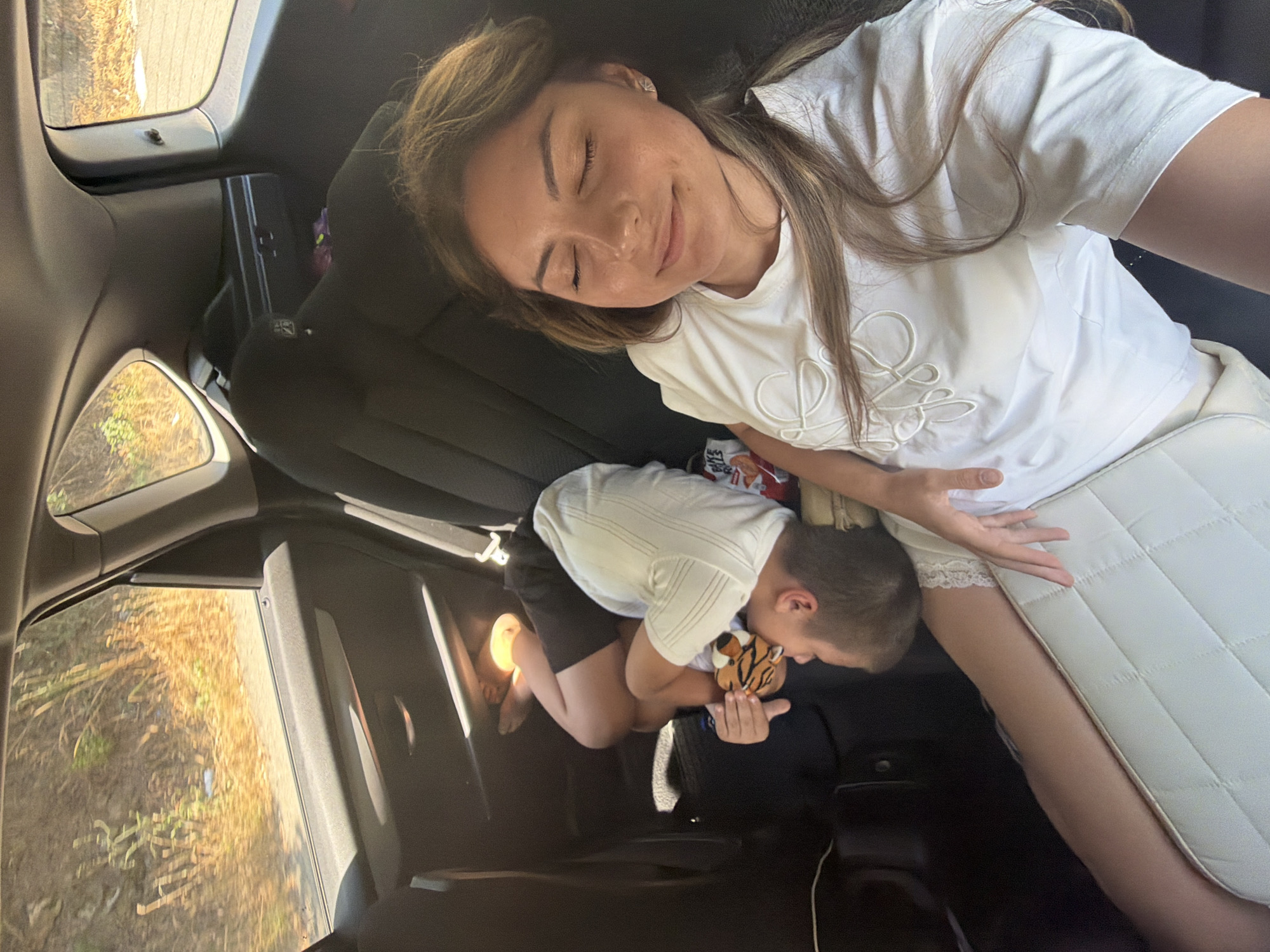 A woman is in the front passenger seat of a car, with her eyes closed and a slight smile. To her left, two children are asleep in the back seat. One child is lying on their side, hugging a stuffed tiger toy. The other child is curled up beside them. The car appears to be traveling on a road surrounded by dry, grassy vegetation. The lighting suggests it is daytime, and the sun is casting shadows through the windows. The interior of the car is dark, with black upholstery.
