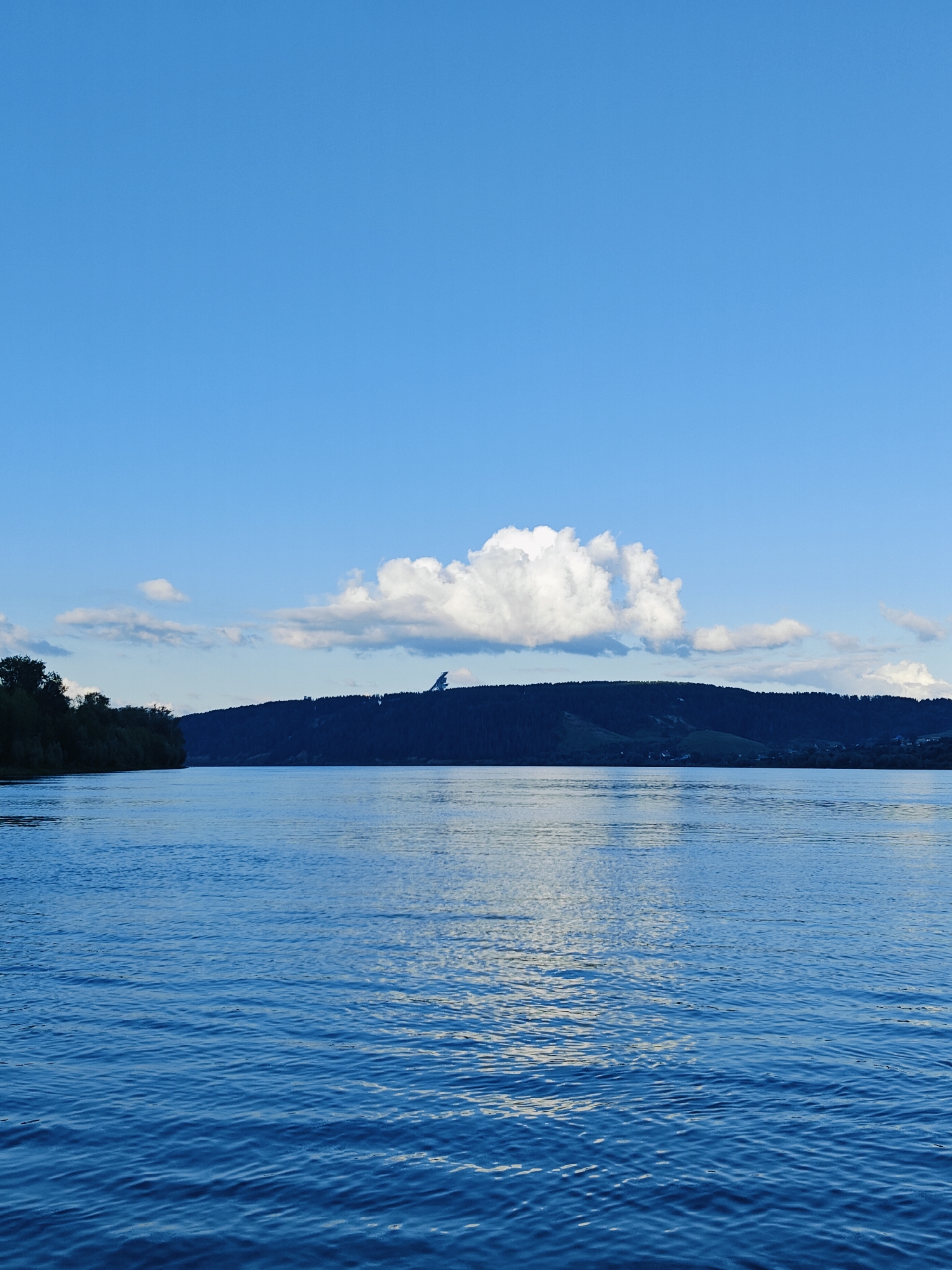 This is a landscape photograph of a wide, calm body of water, likely a river, under a bright blue sky with scattered white clouds. The water occupies the lower half of the frame and exhibits gentle ripples reflecting the sky.
In the background, a dark, forested hill or mountain range stretches across the horizon. The trees are dense and appear as a dark silhouette against the sky. A prominent, distinctive modern structure, possibly a ski jump or similar architectural feature, stands out on the crest of the hill. The structure is silhouetted and appears angular.
To the left of the frame, a line of trees borders the water, suggesting the edge of a wooded area. The sky is a clear, vibrant blue, with one large, fluffy white cloud directly above the hill. The overall lighting suggests it is daytime, likely late afternoon or early evening given the soft light and the presence of shadows on the hill. The scene conveys a sense of tranquility and natural beauty.
The location context provided is Ol'khovskoye, Russia. No text is visible in the image.