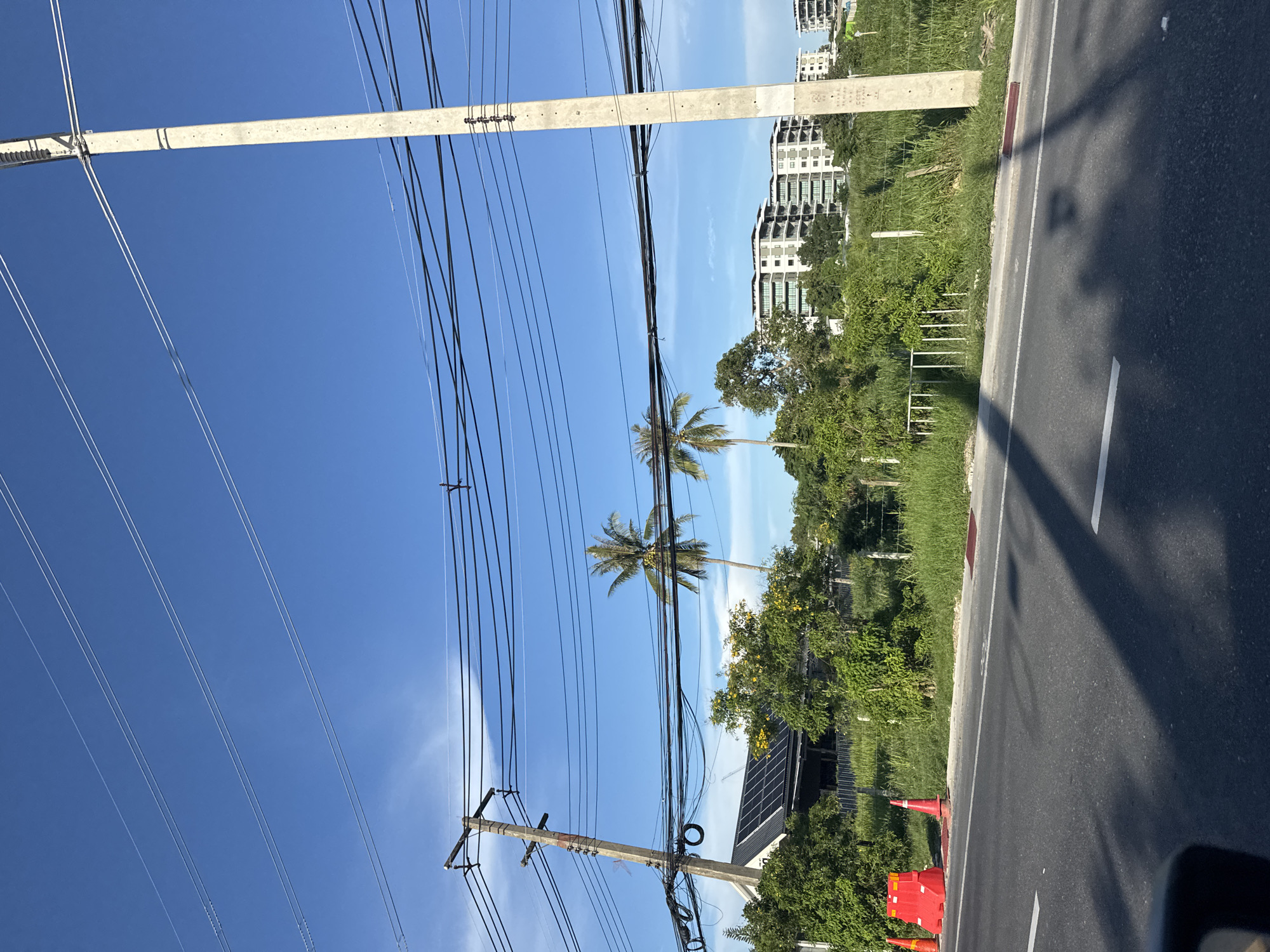 The image depicts a roadside scene with a clear blue sky and a bright sun, casting shadows on the asphalt road in the foreground. The road is marked with white lines, and a few orange traffic cones and a red construction barrier are visible on the right side of the road.
In the middle ground, a patch of green grass is bordered by a wire fence. Behind the fence, a cluster of lush green trees and foliage dominates the landscape, with two prominent palm trees standing tall. On the left side of the image, a house with solar panels on its roof is partially visible. Further in the background, several modern apartment buildings with balconies are situated, suggesting an urban or suburban setting.
Several utility poles with a complex network of overhead wires are scattered throughout the scene, stretching across the frame and partially obscuring the view of the sky and the buildings. The overall impression is one of a somewhat undeveloped area with modern residential structures encroaching on the natural landscape, marked by the presence of essential infrastructure like power lines and traffic management tools. The scene appears to be an outdoor environment, likely during the daytime given the strong sunlight and shadows.