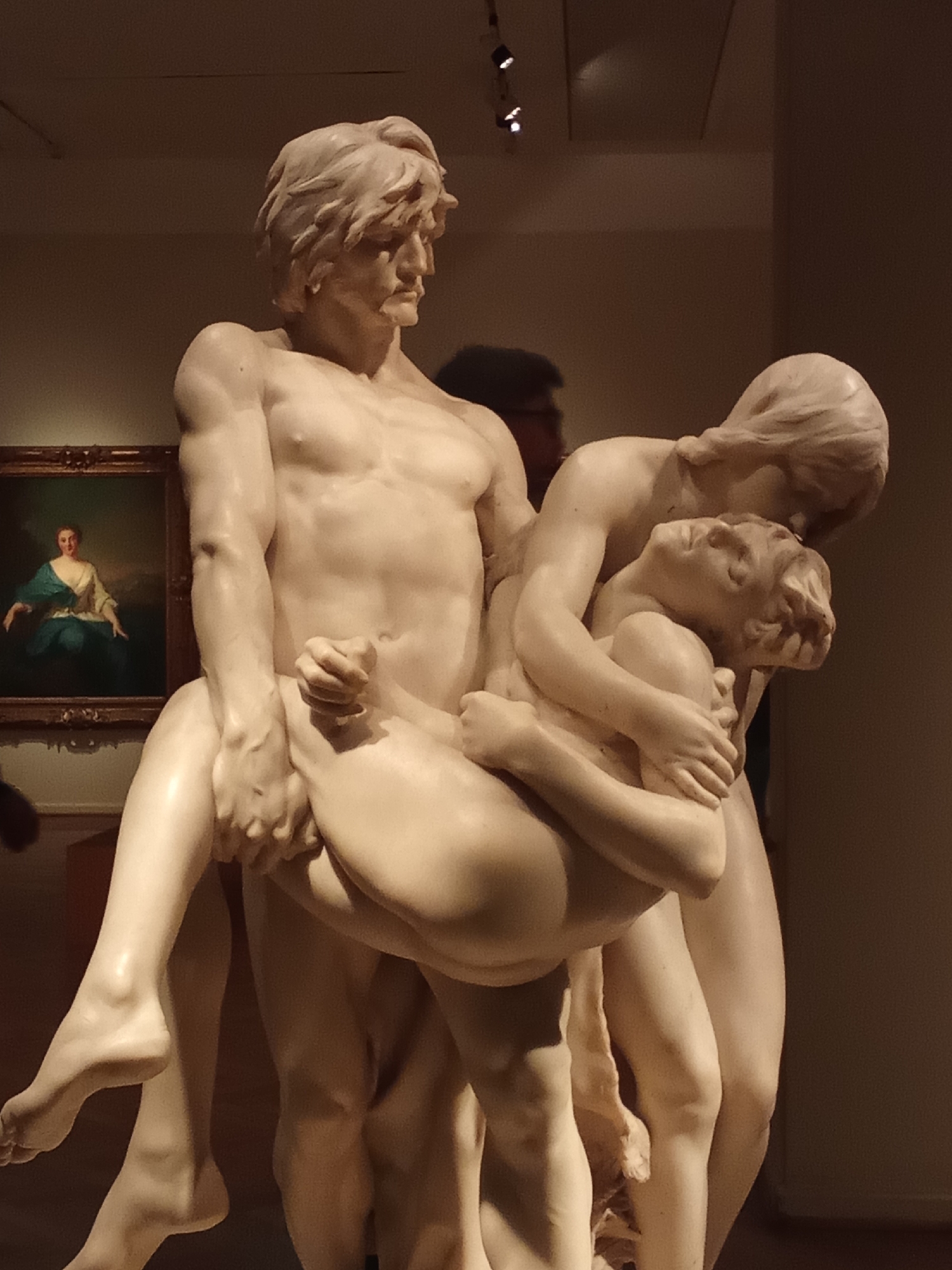 The image depicts a marble sculpture of three nude figures, set in a dimly lit museum gallery. In the foreground, a muscular male figure, depicted from the chest down, carries a reclining female figure across his shoulders. Her head rests on his left shoulder, and her body is draped across his back. A third, smaller female figure is cradled in the arms of the male figure. Her head is turned away from the viewer, and her arms are wrapped around his neck. The sculpture is highly detailed, with realistic musculature and flowing drapery.
In the background, a framed oil painting hangs on a wall. The painting, which is much smaller than the sculpture, depicts a seated woman in historical attire, with a blue garment draped over her shoulders. The overall impression is one of dramatic tension and emotional intensity. The lighting in the gallery is focused on the sculpture, creating strong contrasts of light and shadow. The wall behind the sculpture is a neutral, muted color. The floor is not visible in the frame.
It is not possible to determine the city of Buenos Aires, Argentina from the provided image.