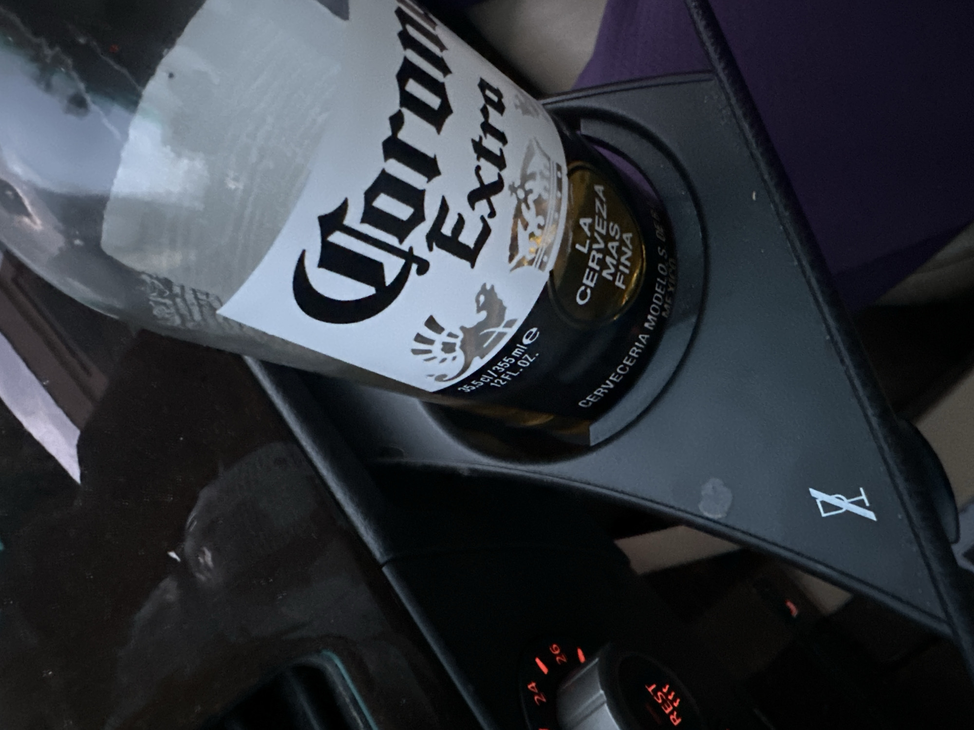The image shows a partially consumed bottle of Corona Extra beer placed in a black plastic cupholder, likely inside a car, given the dashboard details visible in the lower portion of the frame. The bottle's label is clearly visible, detailing the brand and other information. The cupholder has a logo on it. The lighting suggests it might be daytime or an artificially lit interior. The location in Plovdiv, Bulgaria, is not detectable from the image content.