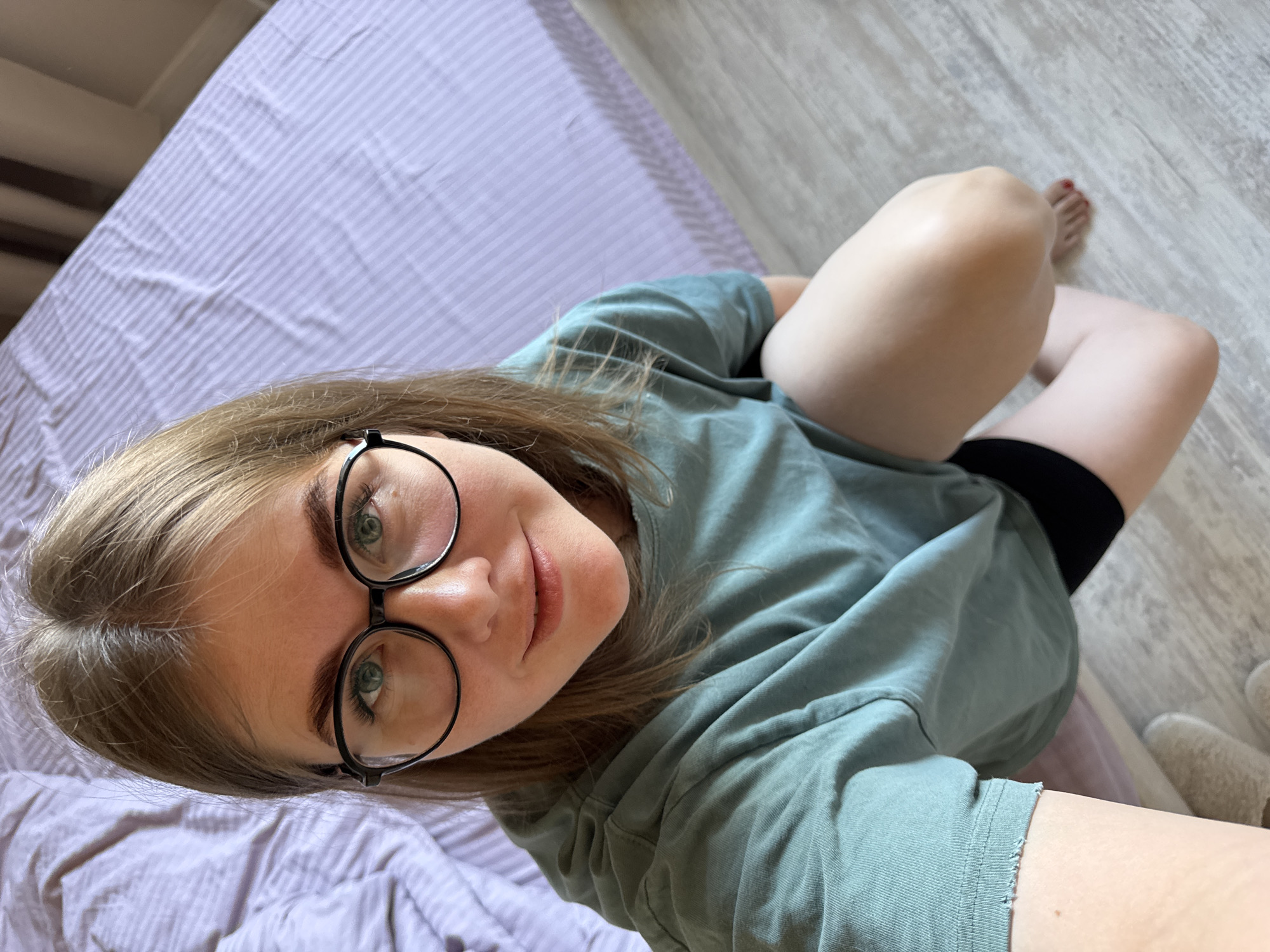 The image depicts a woman with fair skin, light brown hair, and green eyes wearing black-rimmed glasses and a teal t-shirt. She is lying on a bed with a lavender-colored striped bedspread. The woman is angled towards the camera, her face prominent in the foreground. She appears to be looking directly at the viewer. Her legs are bent at the knees. Black shorts are visible beneath the t-shirt. A wooden floor is visible in the background, along with a glimpse of a slipper. There is no verifiable information to suggest that the photo was taken in the city of Ферма, Russia.