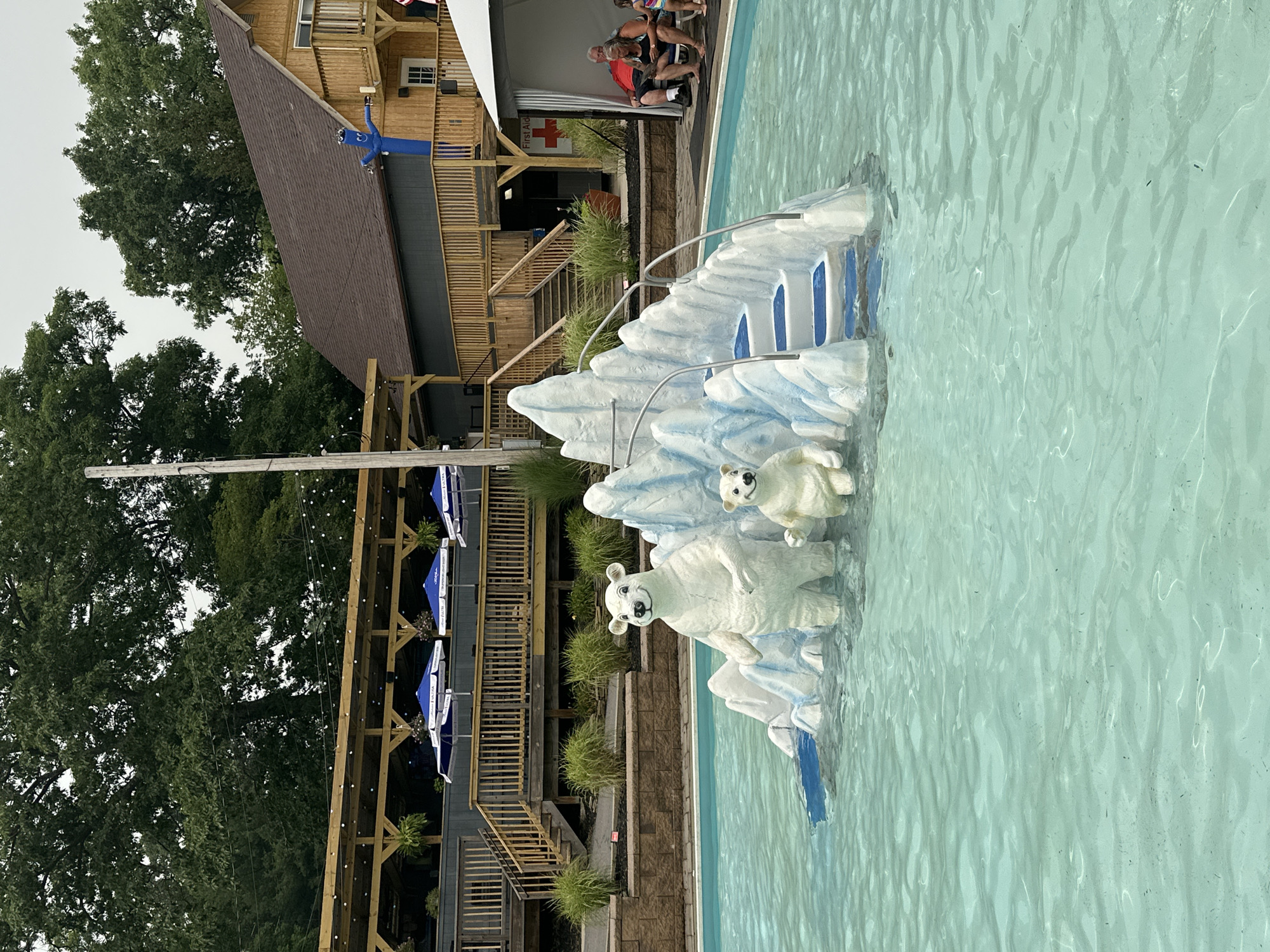 The image shows a swimming pool area at a water park or recreational facility. In the center of the pool, there is a decorative ice floe structure with two polar bear statues. A larger polar bear stands on the left, and a smaller cub is to its right. Steps are integrated into the ice floe, leading up to a platform. In the background, a multi-level wooden structure with balconies and railings is visible, possibly a restaurant or event space. Blue umbrellas are set up on a patio area in front of the wooden building. A tall, blue inflatable tube man stands on a balcony. To the right of the pool, there is a white tent with a red cross symbol on a sign, indicating a first-aid station. A few people are seated on chairs under the tent. The surrounding area is landscaped with grass and bushes, and mature trees are visible in the distance. The sky is overcast. The image appears to have been taken during the daytime in the United States, likely a warm climate given the pool setting, although the specific location in Scranton, USA, is not evident from the visual information alone.