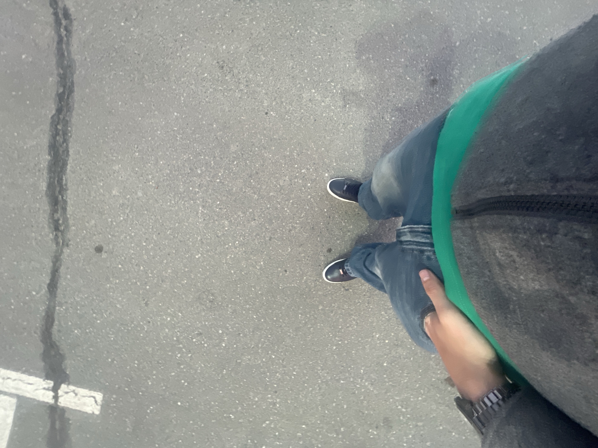 An aerial view shows a person standing on cracked asphalt. The person is wearing blue jeans, black shoes with white soles, and a dark gray jacket with a green stripe. The person's right hand and a watch are visible. The location is unspecified.