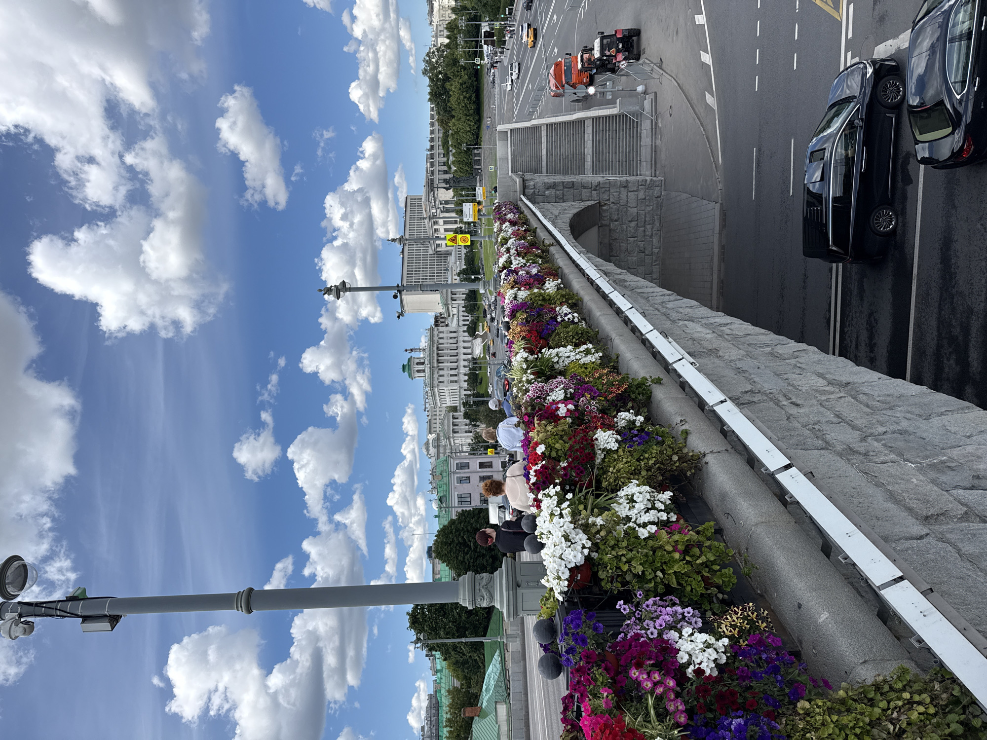 The image depicts a view from a bridge adorned with abundant flower arrangements in Moscow, Russia. In the foreground, a stone bridge railing is covered in a vibrant display of petunias and other flowering plants in various colors, including pink, purple, white, and red. Several people are visible standing on the bridge, looking out at the city or interacting with each other. Beyond the bridge, the cityscape of Moscow unfolds, featuring a variety of architectural styles, including classical and modern buildings. Notable landmarks appear to be present in the distance, with a large, imposing building with a green dome dominating the skyline. A street with traffic, including cars, buses, and construction vehicles like a tractor and a truck, runs beneath the bridge and into the distance. The sky is bright blue with fluffy white clouds, indicating a sunny day. The scene captures a lively urban environment enhanced by decorative floral displays.