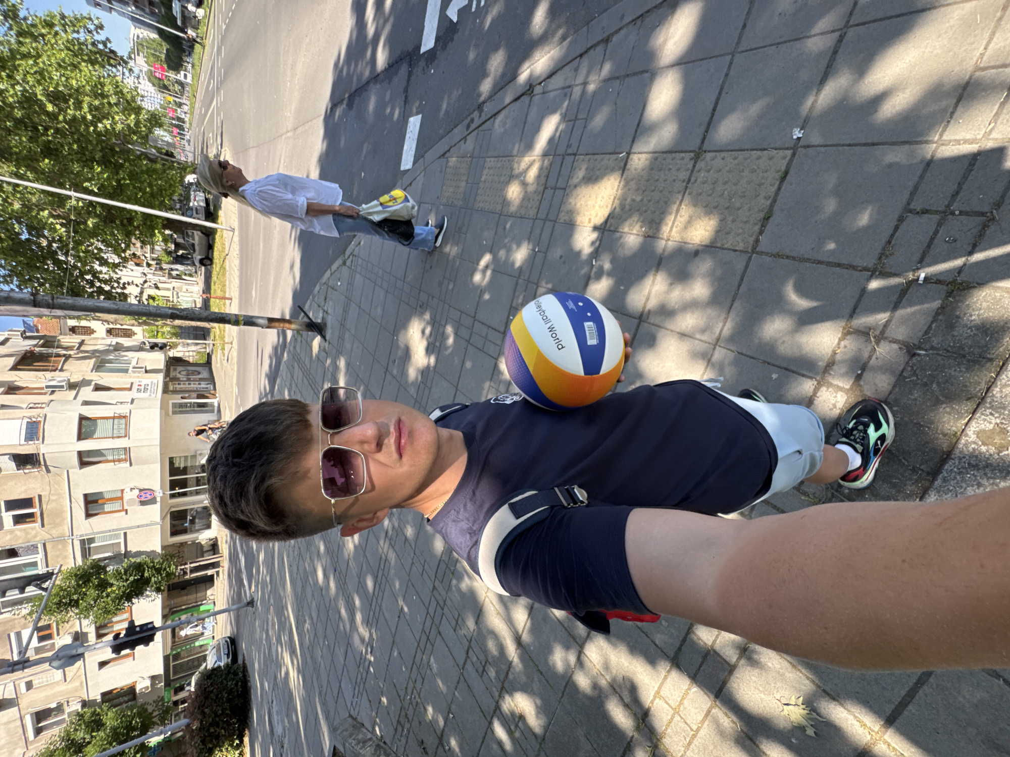A young man wearing sunglasses, a dark blue t-shirt, and light blue shorts is taking a selfie on a sun-drenched city sidewalk. He holds a blue, yellow, and white volleyball. The background shows a multi-story building with shops on the ground floor, a street with traffic, trees, and a woman in a white shirt and jeans crossing the street in the distance. The scene is consistent with a street in Burgas, Bulgaria, a popular summer resort town on the Black Sea coast known for its urban parks and pedestrian zones.