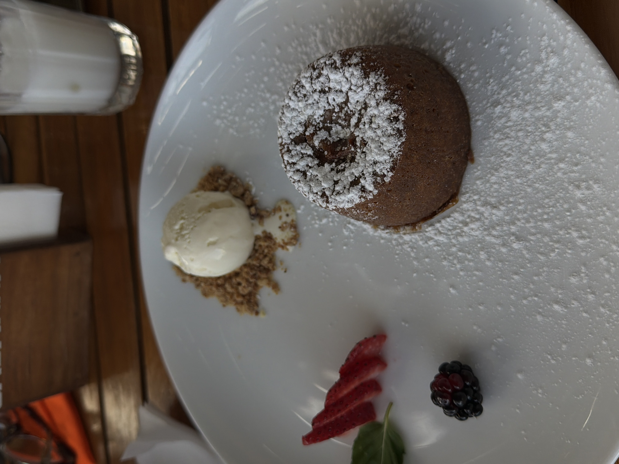 A beautifully plated dessert is the focal point of this image, presented on a white oval dish. The main component is a dark brown, circular cake, resembling a chocolate lava cake, artfully dusted with white powdered sugar. To the left, a scoop of light-colored ice cream, likely vanilla, is served atop a small heap of crushed nuts or granola. Garnishing the lower right portion of the plate are several fanned slices of red strawberry, a single dark blackberry, and a fresh green mint leaf.
The white plate rests on a wooden table, characterized by parallel planks, suggesting a dining setting in a restaurant or cafe. In the upper left corner, parts of a glass containing a white liquid and a small white shaker with a silver cap are visible. The bright, natural lighting indicates it is daytime. There are no people visible, nor any discernible text. This appealing dessert is prepared for consumption, likely in a culinary establishment in Puebla, Mexico.