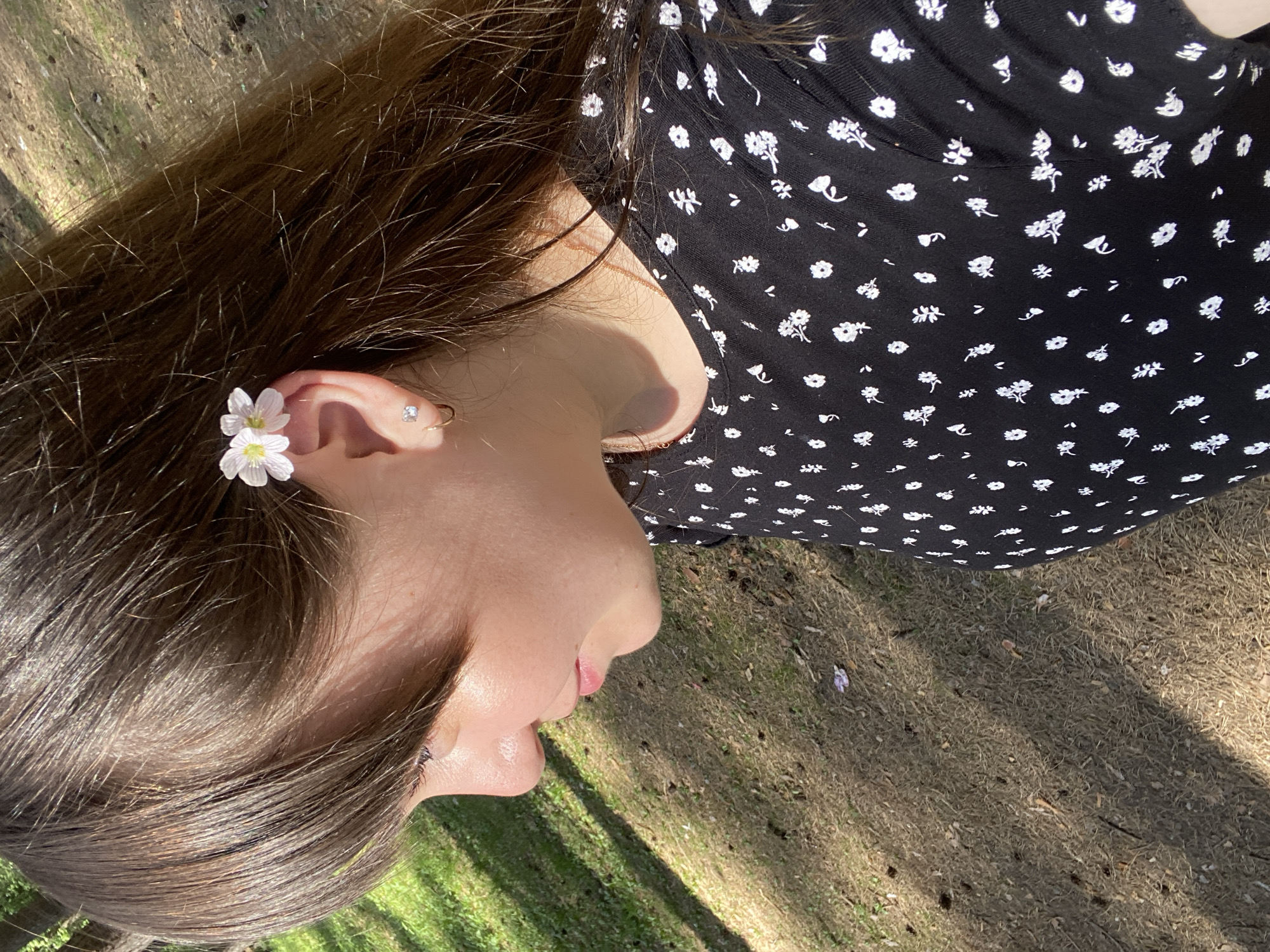 The image depicts a person lying on the ground. They have dark brown hair and are wearing a black dress with small white floral patterns. A small white flower is tucked behind their ear. The person is lying on an area with a mix of dry grass, leaves, and possibly bare soil. Sunlight filters through, creating shadows on the ground. No other people or specific objects are visible in detail. The location in the city of Votkinsk, Russia, cannot be determined from the visual information provided.