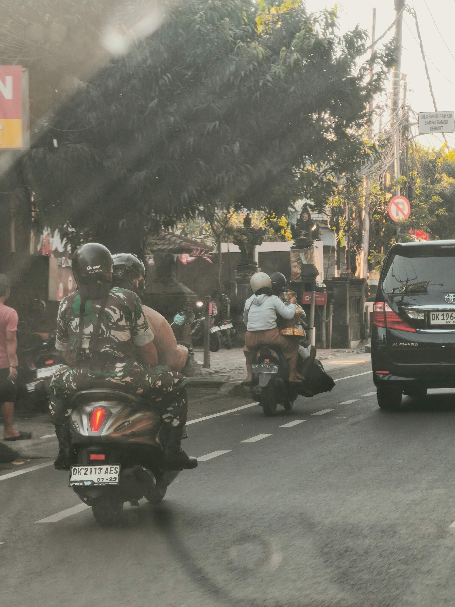 A street scene in Denpasar, Indonesia shows a motorcycle carrying two individuals, likely a man and a woman, from behind. The rider, wearing a camouflage uniform and a black helmet with "HONDA" written on it, appears to be male. The passenger, wearing a light-colored hoodie and brown shorts, is seated behind the rider. The motorcycle has a license plate that reads "DK 2113 AES" and appears to be a Yamaha. To the right, a black Toyota Alphard SUV is visible with the license plate "DK 1964". Further down the street, another motorcycle with two occupants is seen, one wearing a light-colored top and the other a darker top. A "No Parking" sign and a "No Stopping" sign are posted on a utility pole in the background, along with a street sign that reads "DILARANG PARKIR SAMPAI RAMBU BERIKUT". Trees and buildings line the street, and power lines are visible overhead. The lighting suggests it is daytime, possibly late afternoon given the warm tones.
