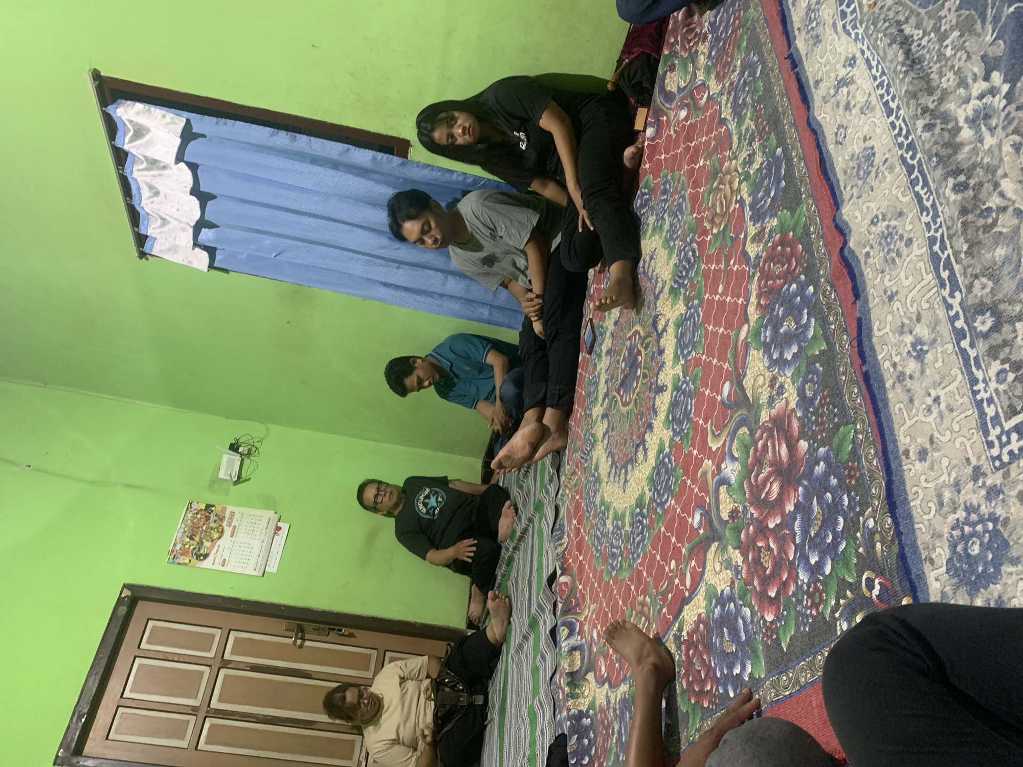 In Dukuh Mojo, Indonesia, a group of six individuals are gathered in a room with lime green walls. The floor is covered with a large, colorful patterned rug featuring floral designs in red, blue, and yellow. A striped mat is laid on top of the rug in the background.
On the left side of the room, a woman with glasses and brown hair tied back sits on the striped mat, wearing a light brown t-shirt and black pants. Next to her, a man wearing glasses, a dark t-shirt with a circular logo, and black pants sits on the mat. To his right, a younger man in a teal t-shirt and black pants is seated. Further to the right, a young man in a grey t-shirt and black pants is sitting, with his eyes closed. Beside him, a young woman with long dark hair is wearing a black t-shirt and black pants. The legs of at least two other individuals are visible in the foreground, also seated on the floor.
The room has a wooden door with panels on the left wall. A calendar hangs on the wall near the door, with text and images. A small white device, possibly a router, is mounted on the wall with wires connected. A window with blue curtains and a decorative valance is on the back wall. The lighting in the room appears to be artificial.