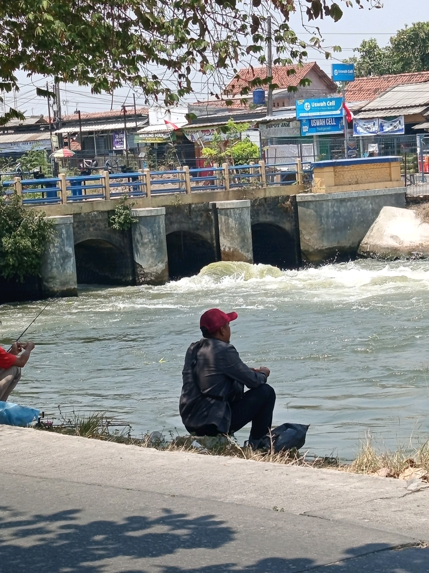 The image depicts a scene along a river in Karawang, Indonesia, with a bridge structure in the background. Two individuals are present. In the foreground, a person wearing a red cap and dark clothing sits facing the river, appearing to be fishing. To the left, another person, partially visible and wearing a red shirt, is also seated and seems to be fishing, holding a fishing rod. The river flows with moderate turbulence, suggesting it's a dam or a point with water flow. The bridge has multiple arches and is constructed of concrete. Along the riverbank, there is vegetation, and a paved walkway runs parallel to the river. In the background, buildings with red-tiled roofs and signs, including one that reads "USWAH CELL," are visible. The overall environment appears to be an urban or semi-urban area with a mix of natural and man-made elements.
