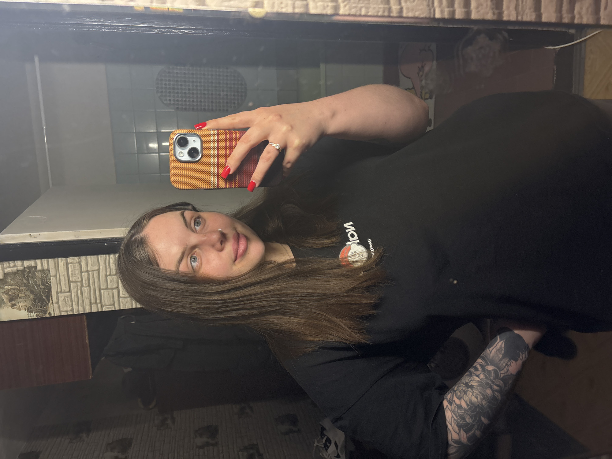 A woman is taking a selfie in a mirror. She is wearing a black T-shirt with a logo on it and has a floral tattoo on her forearm. She has long brown hair, fair skin, and a nose ring. She's holding a cell phone in her right hand, with red nail polish. The case of her cell phone is striped, orange, and yellow. The background includes a tiled bathroom wall and a framed picture, suggesting a bathroom setting. The image was possibly taken in Chornomorskoe, Ukraine.