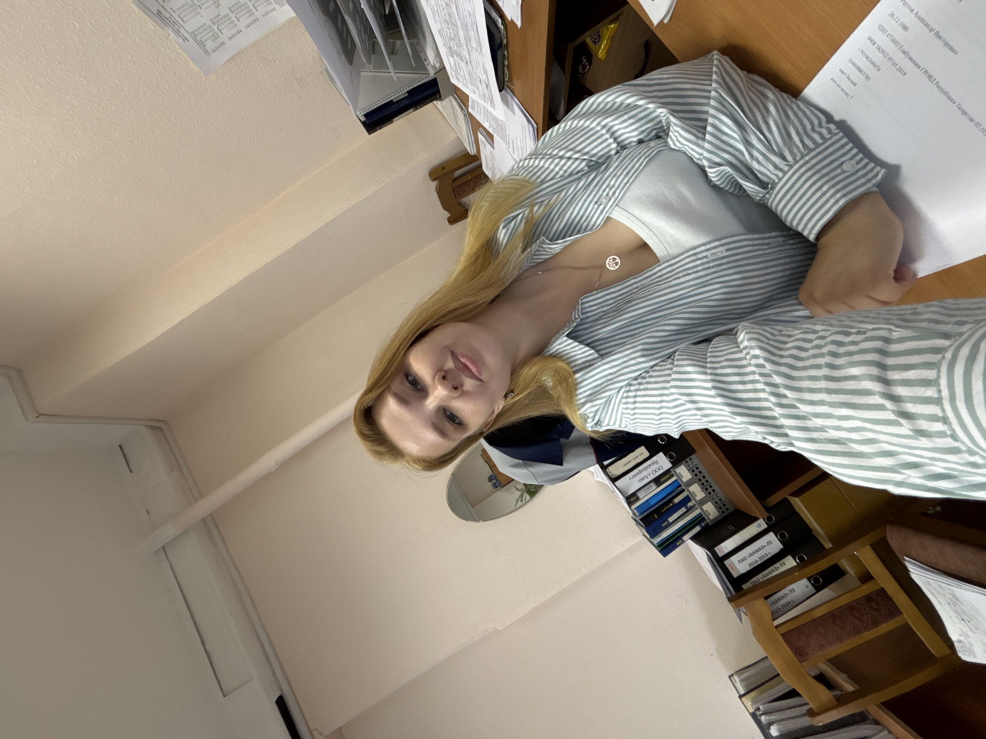 The image depicts a young woman with blonde hair sitting at a desk in an office. She is wearing a white tank top and a light blue and white striped button-down shirt. A silver necklace with a tree pendant is visible on her chest. The desk is cluttered with papers and folders. Behind her, there are several binders on a shelf, a mirror, and a chair. The walls are painted a light peach color, and the ceiling has fluorescent lighting. One document on the desk contains Cyrillic text, suggesting the location is likely in Russia. The context provided about the city of Заволжье, Russia, aligns with this observation. The woman is looking at the camera and smiling, suggesting a casual or informal moment, possibly a selfie. The overall impression is of a work environment, but the selfie suggests a break or a moment of personal connection.