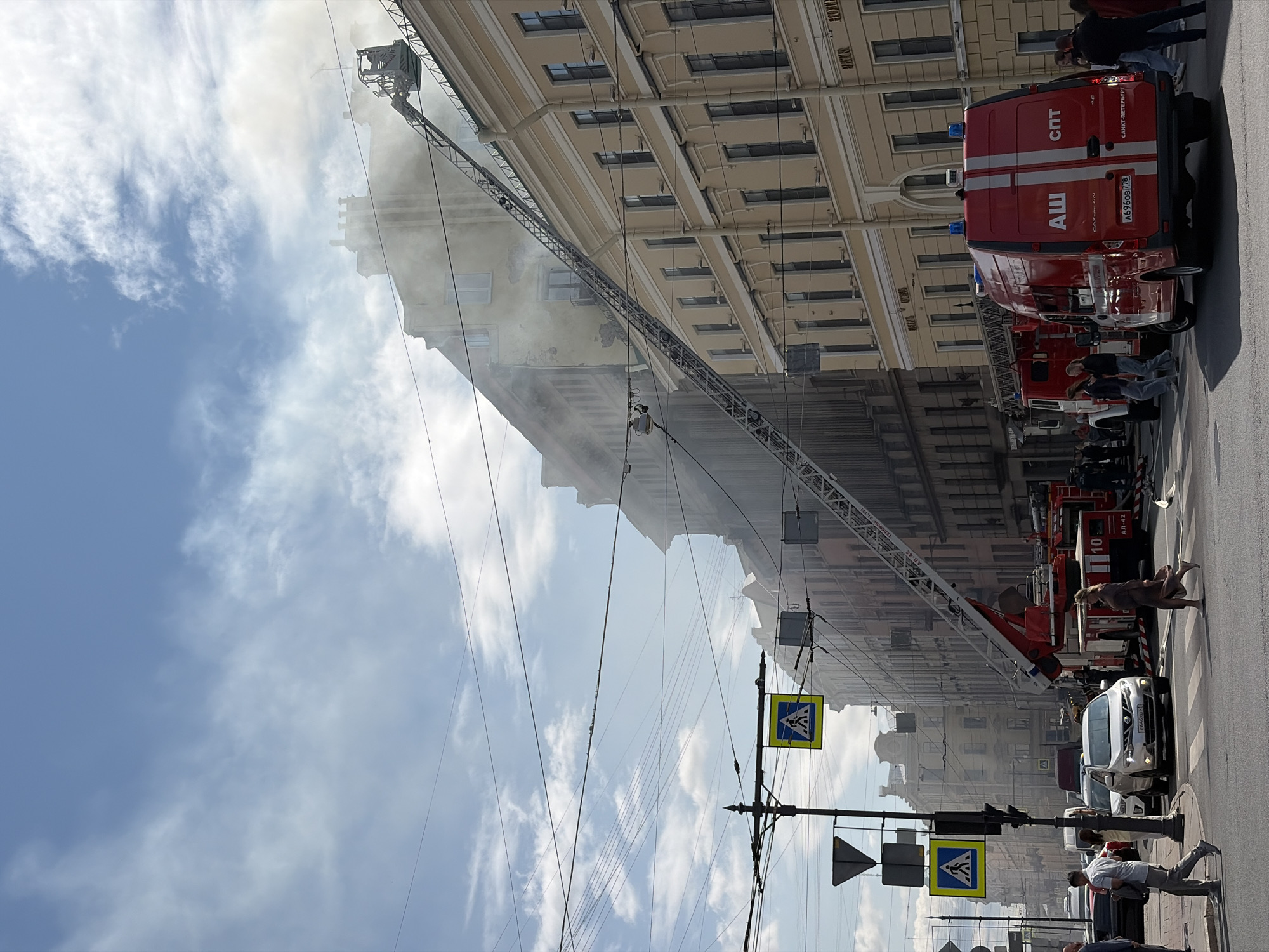 The image depicts a fire scene on a sunny day in Saint Petersburg, Russia. A tall building is engulfed in smoke, with smoke billowing from the upper floors and windows. A fire truck ladder extends to the building, reaching near the source of the smoke. Another red fire truck is parked on the street, with firefighters visible near it. Pedestrians are crossing a street in the foreground, some looking towards the fire. Traffic lights and road signs are present, including a pedestrian crossing sign. The street appears to be in a city, with several multi-story buildings lining it. The sky is blue with scattered clouds. The overall scene suggests an active emergency response to a fire.
