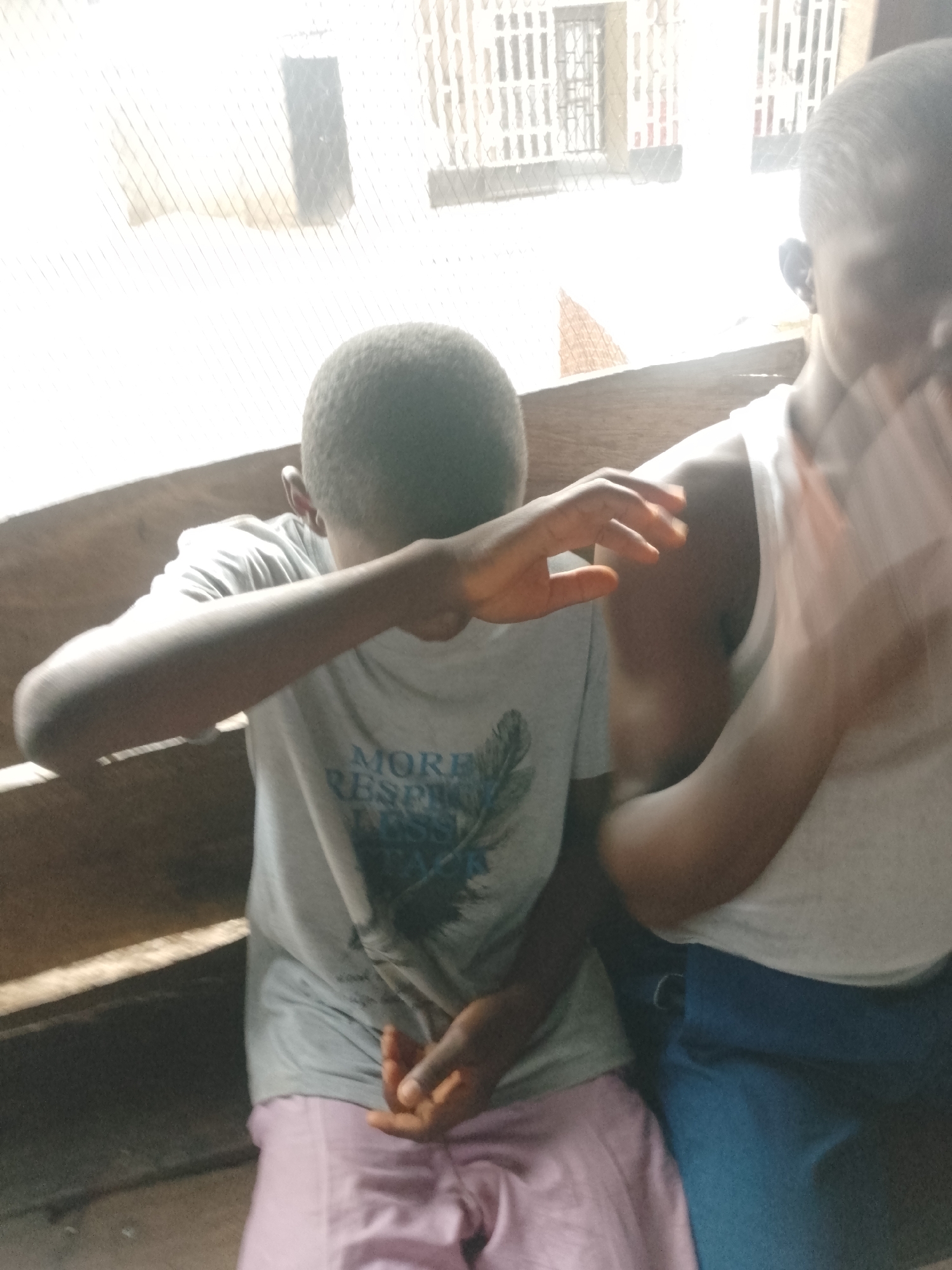 Two children sit next to each other on a wooden bench. The child on the left is wearing a gray t-shirt and pink pants, covering their face with their hand. The child on the right is wearing a white tank top and blue pants, with their arm moving. Behind them is a window with bars. The location is Unknown, Unknown.