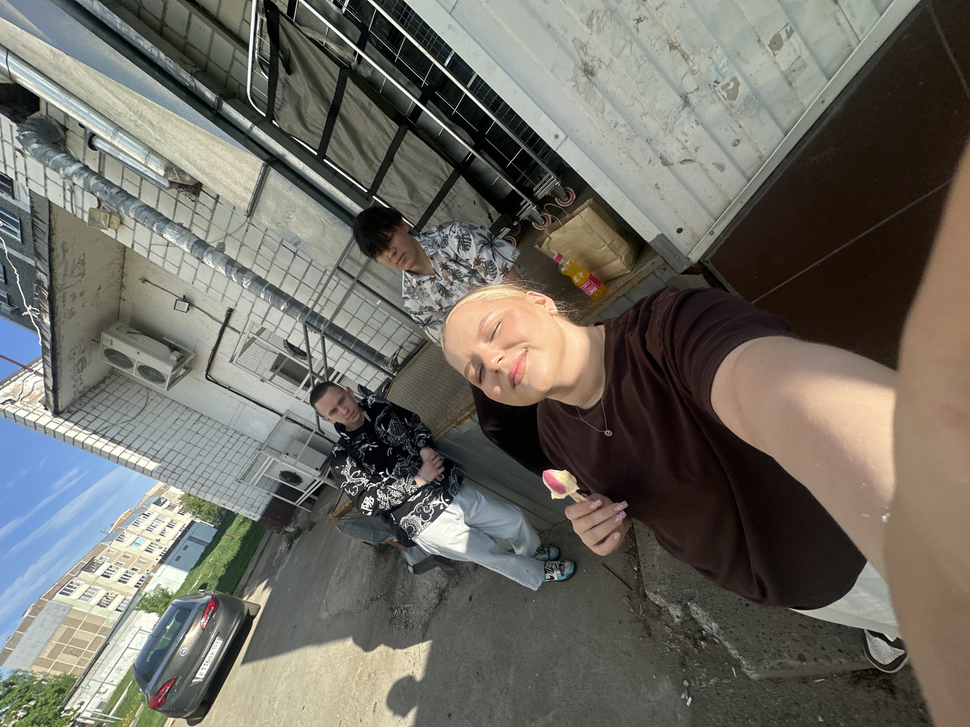 The image, taken outdoors in the city of Chaikovsky, Russia, depicts three individuals in a selfie-style photograph. One person is holding the camera, smiling and holding a flower bud. One individual is wearing a black and white patterned jacket and light-colored pants. Another is wearing a patterned shirt. The environment appears to be an urban setting. A gray car is visible in the background, parked on a paved surface. The background includes residential buildings and greenery. The weather appears sunny.