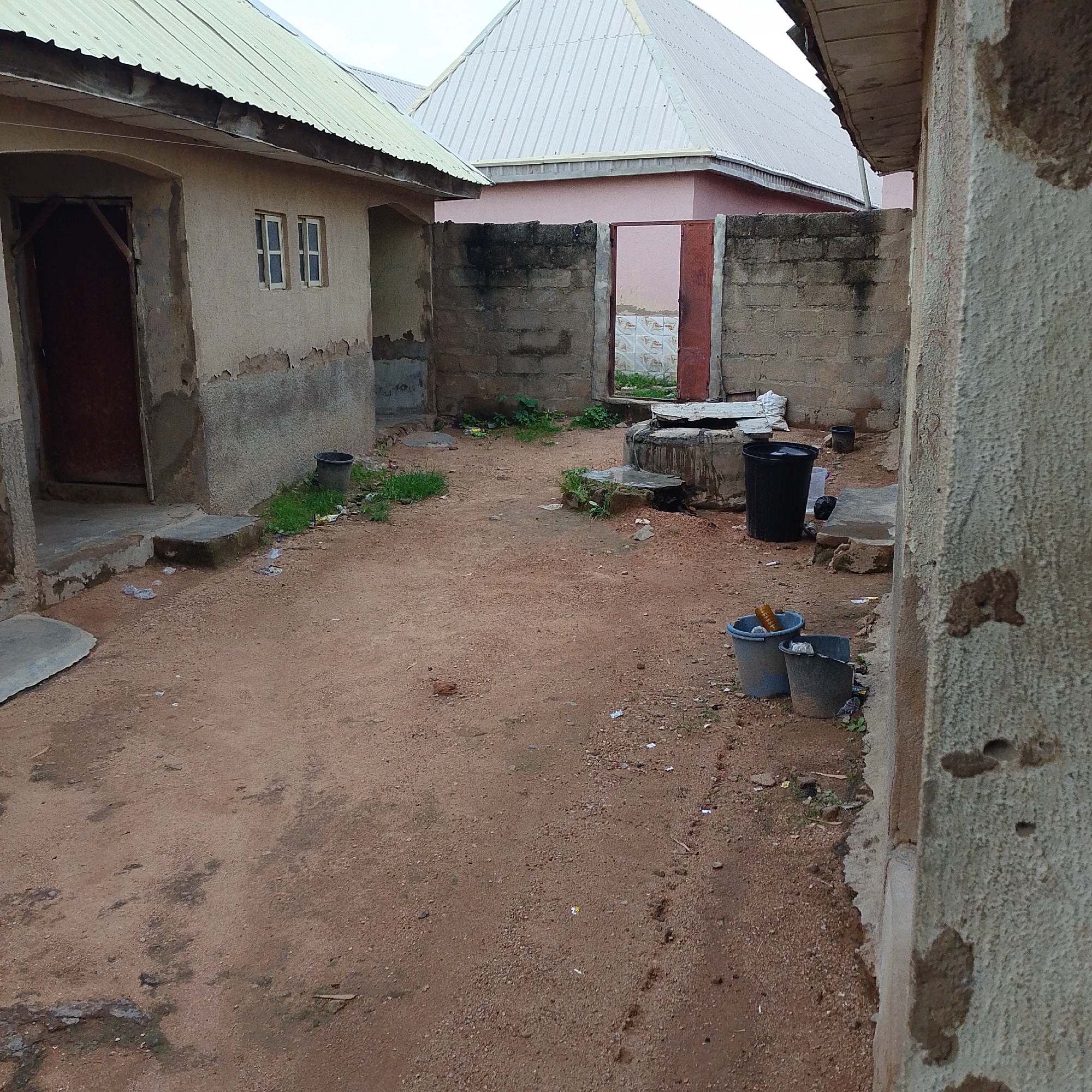 The image depicts an outdoor courtyard area in Jalingo, Nigeria, characterized by its rustic and somewhat dilapidated state. The ground is covered in reddish-brown soil and gravel, with scattered debris including small pieces of white paper and other detritus. Several structures are visible, primarily buildings with rough, textured walls and corrugated metal roofs.
To the left, a building features a light-colored facade with peeling paint near the base, a dark-colored wooden door, and two small, square windows with white frames. The roof of this building is light green.
In the background, a more prominent building with a steeply pitched, white corrugated metal roof is visible, positioned behind a low, dark-colored brick wall. This wall has an opening with a rusty metal gate, beyond which can be seen more of the pink-colored building and a tiled wall inside.
Various objects are scattered around the courtyard. A large black bucket sits near the left building, and further into the courtyard, closer to the brick wall, are several more buckets. One is a light blue bucket containing what appears to be an orange bottle and a plastic bottle. Next to it is a dark grey bucket. A concrete well or cistern with a rough, circular edge is situated in the middle-right of the frame, appearing somewhat aged. Another large black bucket is positioned behind this well.
Vegetation is sparse, with small patches of green grass growing in various locations, particularly around the base of structures and near the well. The overall impression is of a functional, lived-in space that may be experiencing some neglect. The lighting suggests it is daytime, possibly overcast or late afternoon.