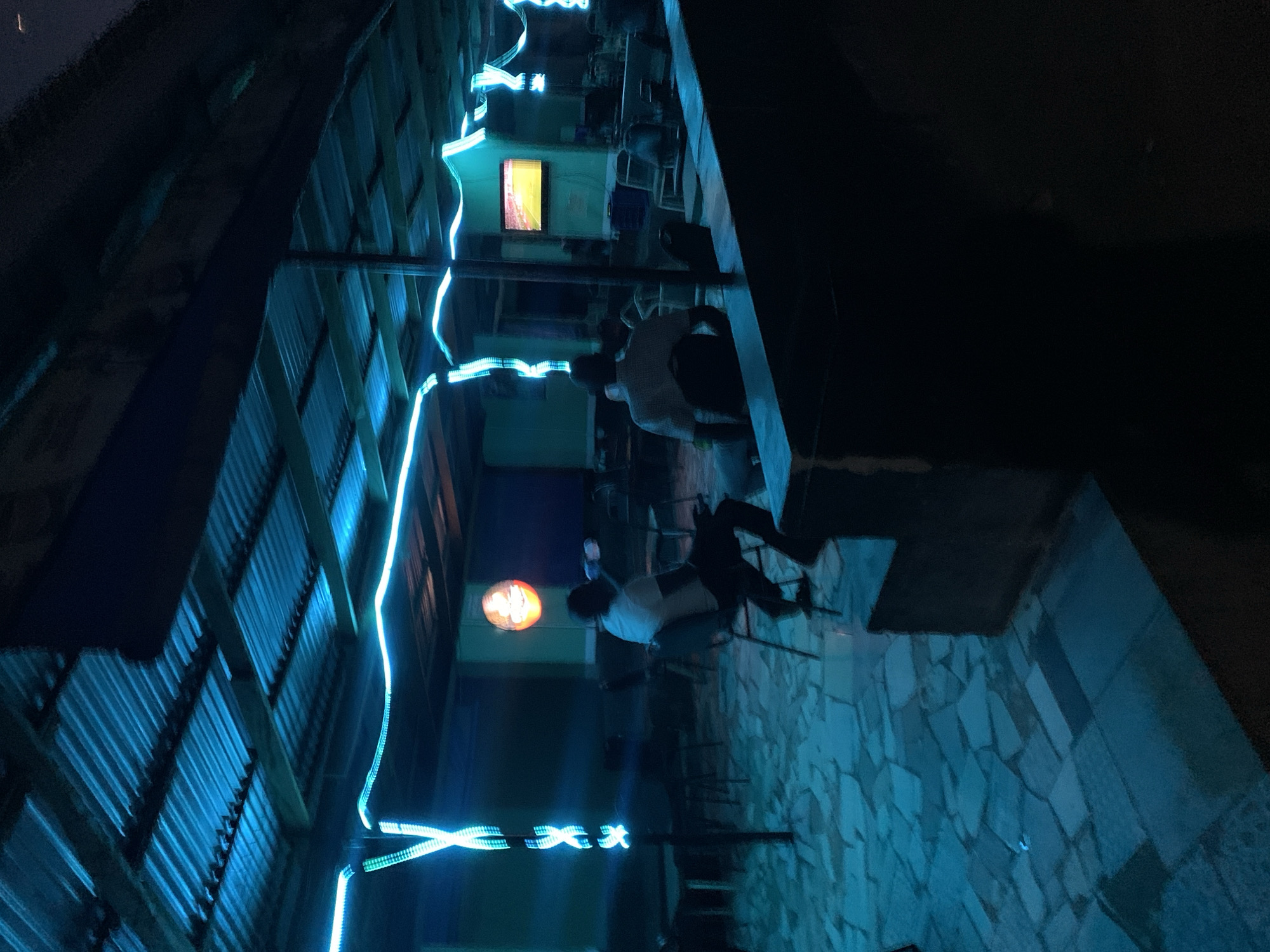 The image depicts an outdoor or semi-outdoor night scene, likely a bar, cafe, or lounge in Kabong, Nigeria. The setting is primarily illuminated by artificial lights, creating a cool, blue ambiance.
The ground is paved with irregular, light-toned flagstones or cobblestones. A prominent feature is a corrugated metal wall or structure, running vertically along the left side of the frame, which is adorned with bright, electric blue neon-like lights. These lights are arranged in wavy lines and decorative 'X' shapes.
At least two individuals are present in the mid-ground. One person, wearing a light-colored shirt and dark trousers, is seated at a table or near a counter, facing away from the camera. Another individual, also in a light shirt, is seated further back and partially obscured. They appear to be engaged in social activity or relaxing in this evening environment.
In the background, various structures can be seen. A distinctive round sign glows with an orange light, while a rectangular window emits a warm yellow-orange light, providing a color contrast to the dominant blues. The overall atmosphere is dark, with the artificial lighting being the primary source of illumination, suggesting a late-night setting. No discernible text is visible.