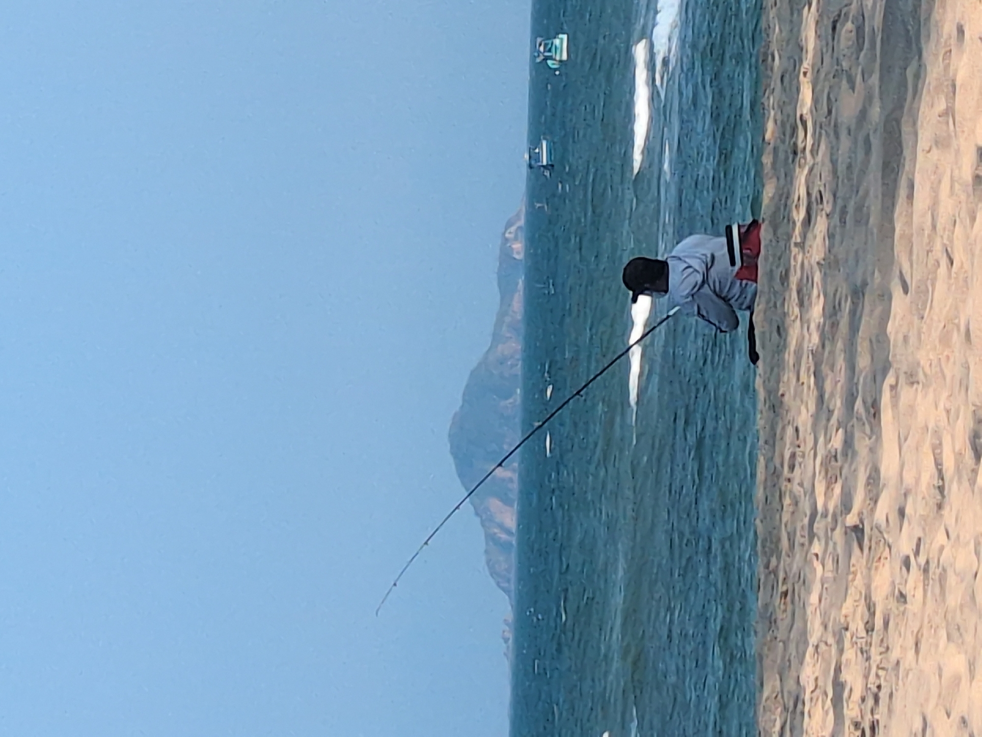 The image depicts a man fishing on a beach, likely in the city of South Quy Nhon Ward, Vietnam. The man is seen bending over, holding a fishing rod. The scene features a sandy beach that appears to meet the ocean. In the background, there is a mountain range and a clear sky. There are boats in the water and waves breaking on the beach.