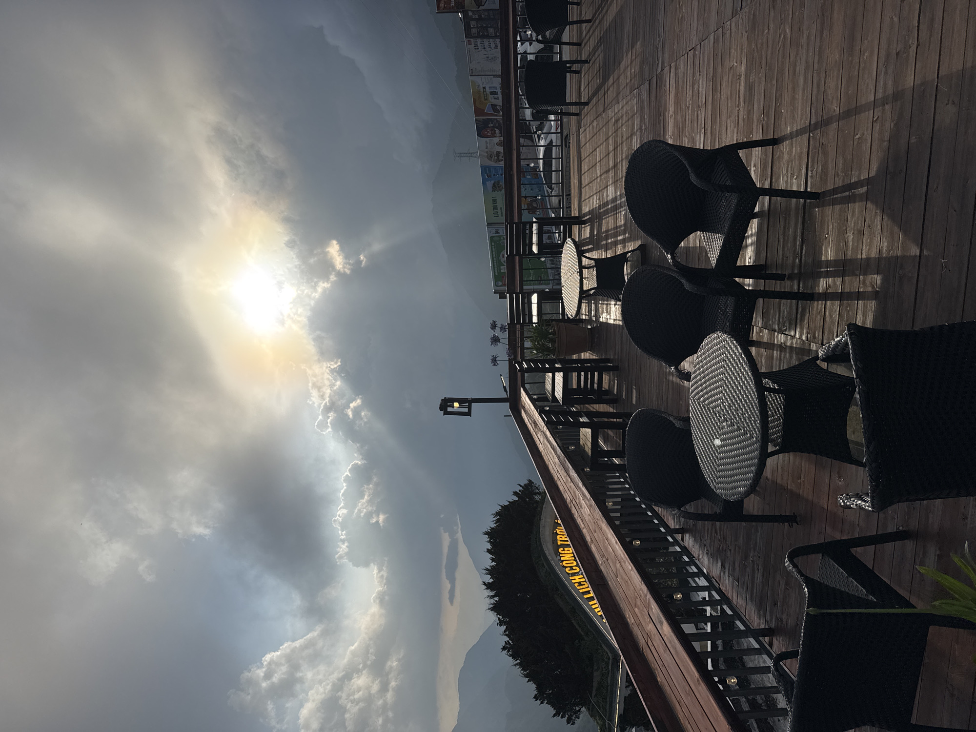 The image depicts an outdoor seating area, likely a deck or patio, in Binh Lu, Vietnam. Woven black patio furniture, consisting of tables and chairs, is arranged on wooden planks. A railing borders the deck, beyond which a view of distant mountains is visible, partially obscured by clouds and the setting sun. Sunlight streams through the clouds. In the background, signage with the words "LICH CONG THANH" in yellow text can be seen.