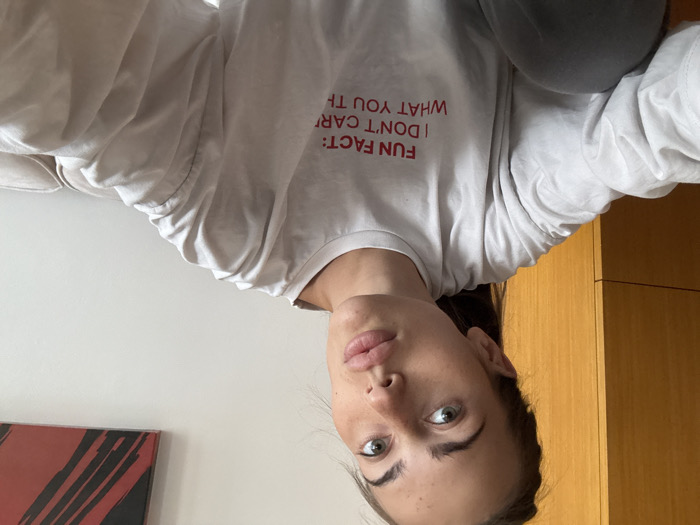 The image depicts an indoor scene in Warsaw, Poland, featuring a female subject capturing a selfie. The woman, with light skin, prominent freckles, light-colored eyes, and dark hair pulled back, looks directly at the camera. She is dressed in a light-toned, long-sleeved shirt emblazoned with the red text 'FUN FACT: I DON'T CARE WHAT YOU THINK' across the chest, and gray trousers, partially visible. The immediate background to the subject's left is a light brown wooden wardrobe or cabinet. To her right, a plain white wall holds an abstract artwork characterized by red and black brushstrokes in the upper corner. A portion of a light-colored, upholstered sofa or chair is visible in the lower right foreground. The environment is uniformly lit, consistent with an interior domestic or private space.