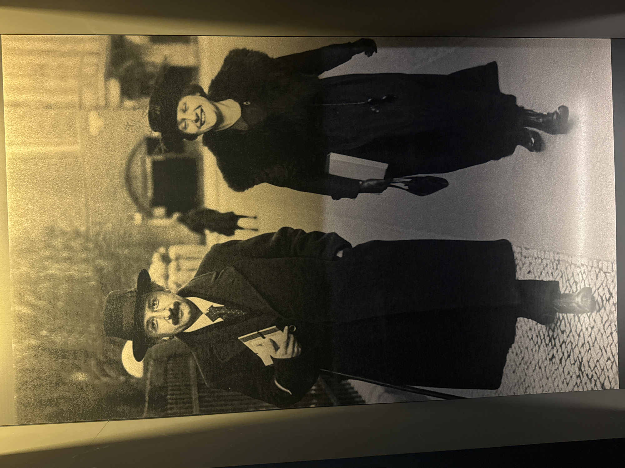 In a sepia-toned, full-length photograph, Albert Einstein and his wife, Mileva Marić, are captured walking on a cobblestone street. Einstein, on the left, wears a dark overcoat, a fedora, and a suit with a patterned tie. He carries a book in his left arm. Mileva, on the right, is smiling and wears a dark coat with a fur collar, a cloche hat, and gloves. She carries a book in her left hand and a handbag hangs from her right wrist. Behind them, blurred figures and architectural elements suggest an urban setting, possibly the city of Bern, Switzerland, where Einstein developed his theory of relativity. The lighting creates a soft, ethereal glow, emphasizing the historical and somewhat nostalgic feel of the image. The perspective is eye-level, as if the viewer is walking alongside them.