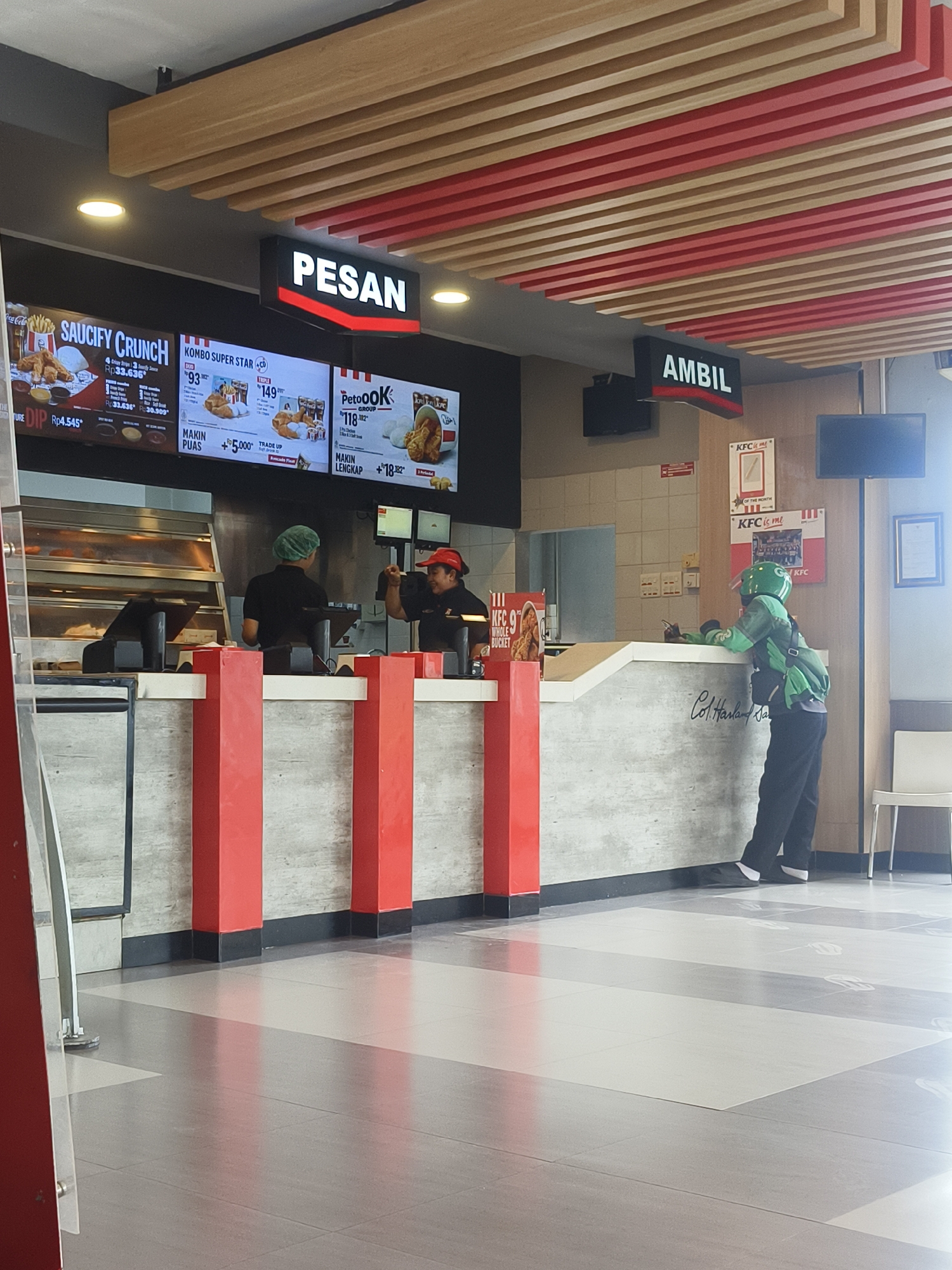 The image depicts the interior of a KFC restaurant in Denpasar, Indonesia. Two employees are visible behind the counter, one wearing a green hairnet and the other a red KFC uniform and hat. A delivery person, dressed in a green uniform with a helmet, is standing at the counter, seemingly waiting for an order. Digital menu boards display various KFC meal options and prices, including "SAUCIFY CRUNCH," "KOMBO SUPER STAR," and "PetoOK GROUP." The restaurant's interior features a modern design with a wood-paneled ceiling and red and beige accents. The floor is tiled in a pattern of grey and white squares, with some foot-shaped markings indicating social distancing guidelines. The overall scene suggests a typical day at a fast-food establishment, with staff serving customers and a delivery driver awaiting a pickup.
