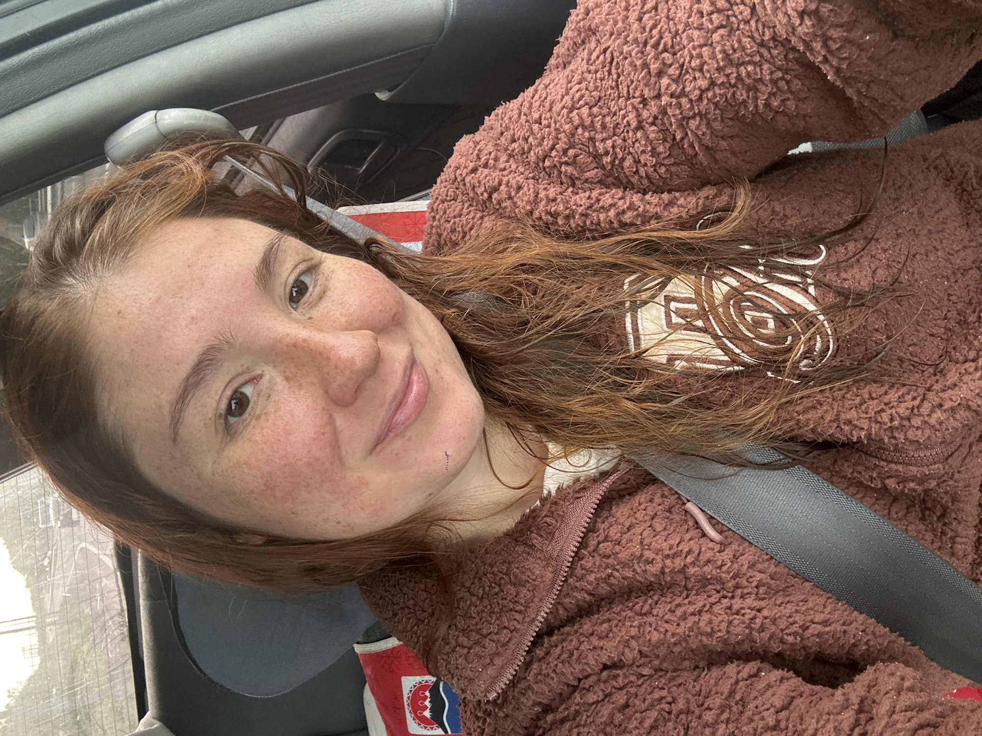 The image is a close-up selfie taken from inside a vehicle, likely a car. A young woman with fair skin, freckles, and auburn hair is the primary subject. She is wearing a brown, fuzzy zip-up jacket over a white top, and a grey seatbelt is visible across her chest. The background shows a blurry view through the car window, suggesting an outdoor environment with some structures and vegetation, possibly indicating a semi-urban or suburban setting. The lighting suggests it is daytime. The location is specified as the city of Petropavlovsk-Kamchatsky, Russia, though this information cannot be visually confirmed from the image itself.