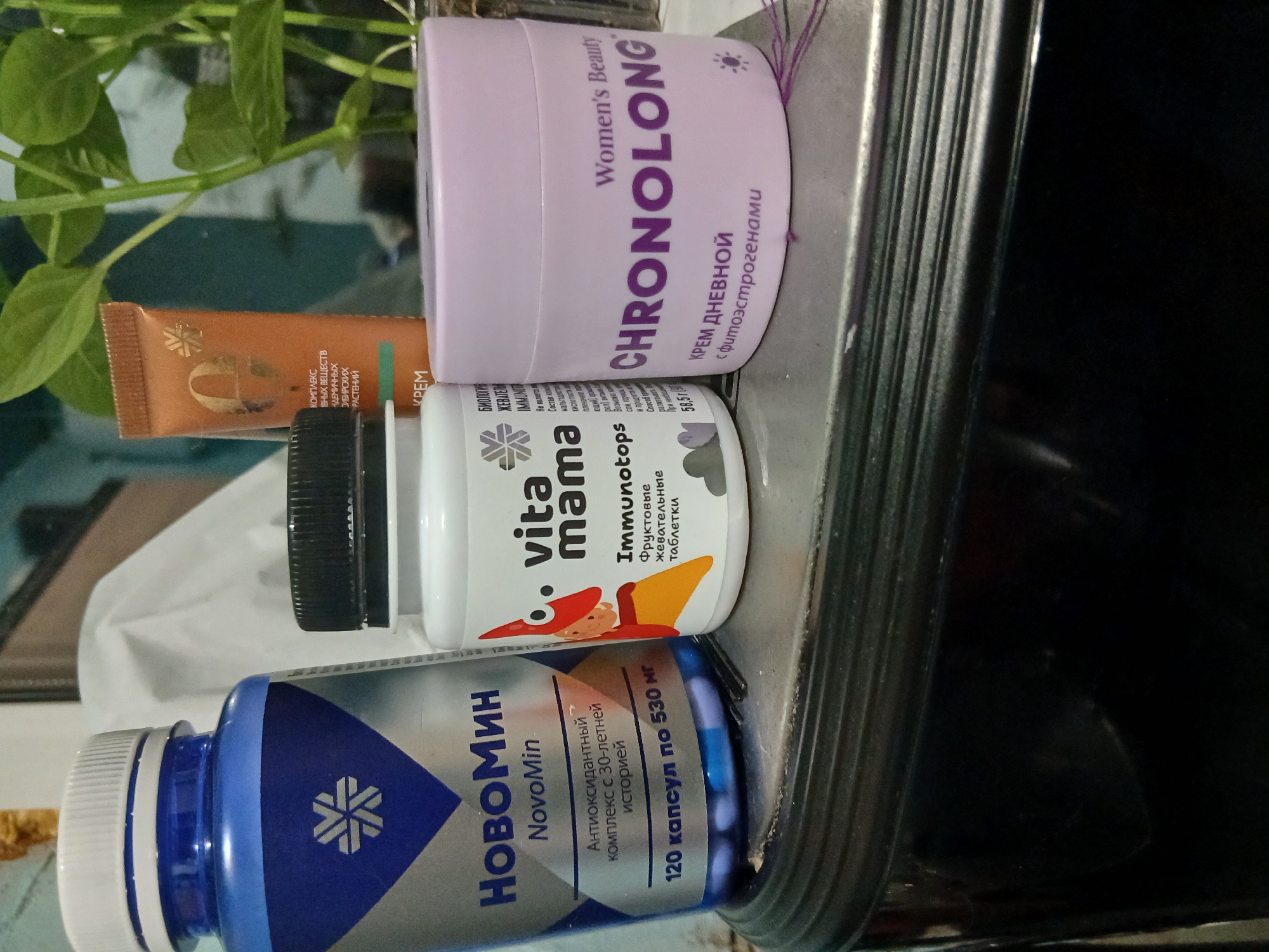 Here's a detailed description of the image:
**Scene Overview:** The image presents a close-up shot of several health and beauty products arranged on what appears to be a metallic or reflective surface, possibly a shelf or tray.
**Objects:**
* A blue and white bottle labeled "НовоМин NovoMin" (Novomin). It is described as an antioxidant complex. The bottle states "120 capsules of 530 mg".
* A white and green bottle labeled "vita mama Immunotops". The label includes a cartoon graphic.
* A light purple container of "CHRONOLONG" women's beauty day cream.
* A small, brown tube of skin cream is also present.
**Environment:** The presence of greenery partially visible in the upper left corner suggests an indoor setting, possibly near a plant. The reflective surface and hints of dark elements may indicate a modern or utilitarian interior, such as a bathroom or kitchen.
**Location:** Based on the Cyrillic writing on the product labels (НовоМин, vita mama, CHRONOLONG), the image is likely taken in a region where Russian or a closely related language is used. There is no explicit indication of the city of Ильинка in the image itself, so it is impossible to include it.