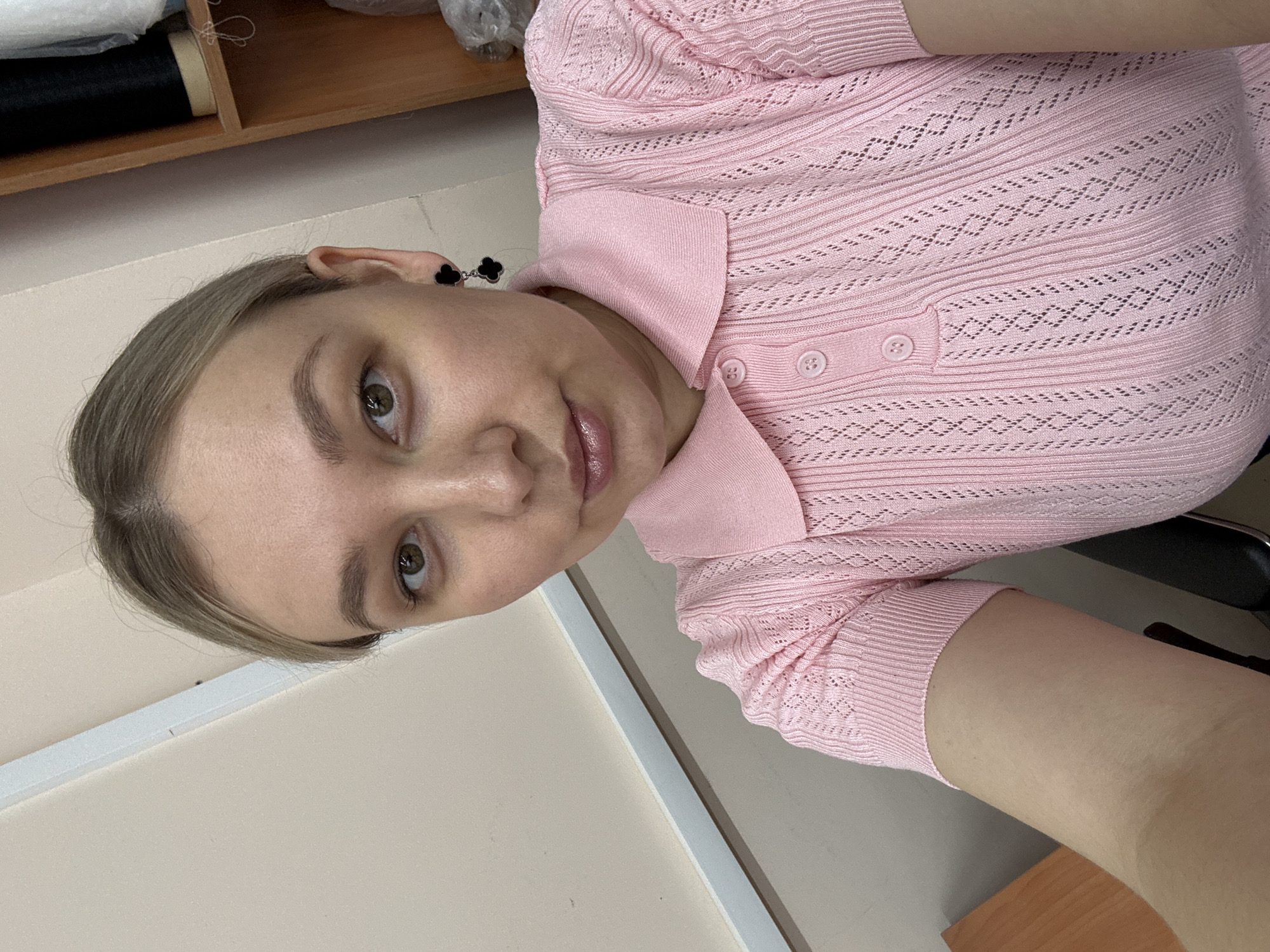 A fair-skinned woman with light brown hair is captured in a close-up photograph. She is wearing a pink, short-sleeved knit top with a collar and two buttons visible. Black, clover-shaped earrings adorn her ears. The background includes a shelf with various items, a white wall, and a desk surface. The location is Chaikovsky, Russia.