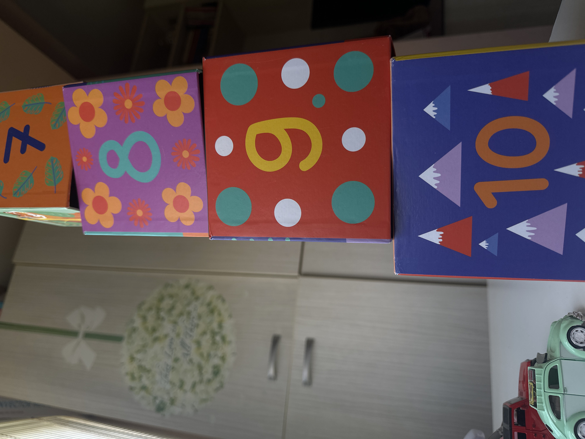 The image shows a stack of four colorful children's blocks. The blocks are stacked vertically and are decorated with numbers and patterns. The top block is orange with a blue number 7 and green leaf patterns. The second block is purple with a teal number 8 and floral designs. The third block is red with a yellow number 9 and scattered green and white polka dots. The bottom block is blue with orange mountains and a large orange number 10. To the left of the blocks, a light green toy car and a red toy car are visible on a white surface. In the background, a blurred wardrobe with two silver handles is visible. The overall scene appears to be indoors, possibly in a child's room or play area, with no discernible elements suggesting a specific location within Sofia, Bulgaria.