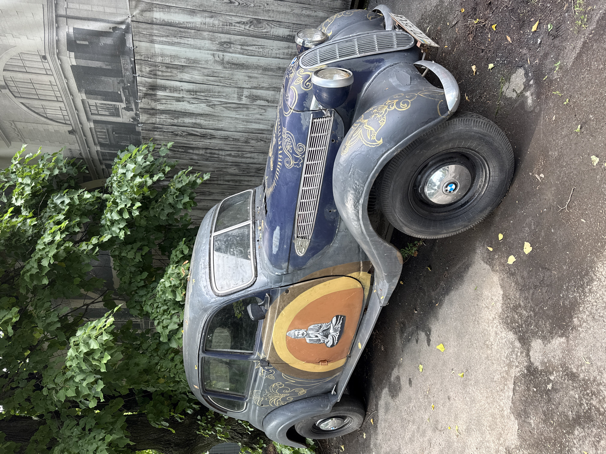 A vintage dark blue and gray BMW automobile, adorned with gold ornamental decals and a circular graphic of a meditating Buddha on the driver's side door, is parked on an asphalt surface in front of a wooden fence and a large tree. The car's headlights and grille are visible. The wooden fence is backed by a large black and white mural depicting an architectural facade. Fallen leaves are scattered on the ground around the vehicle. The scene appears to be located outdoors in Vinnytsia, Ukraine, possibly in an urban setting, given the presence of the mural and the asphalt. There are no people visible in the image.