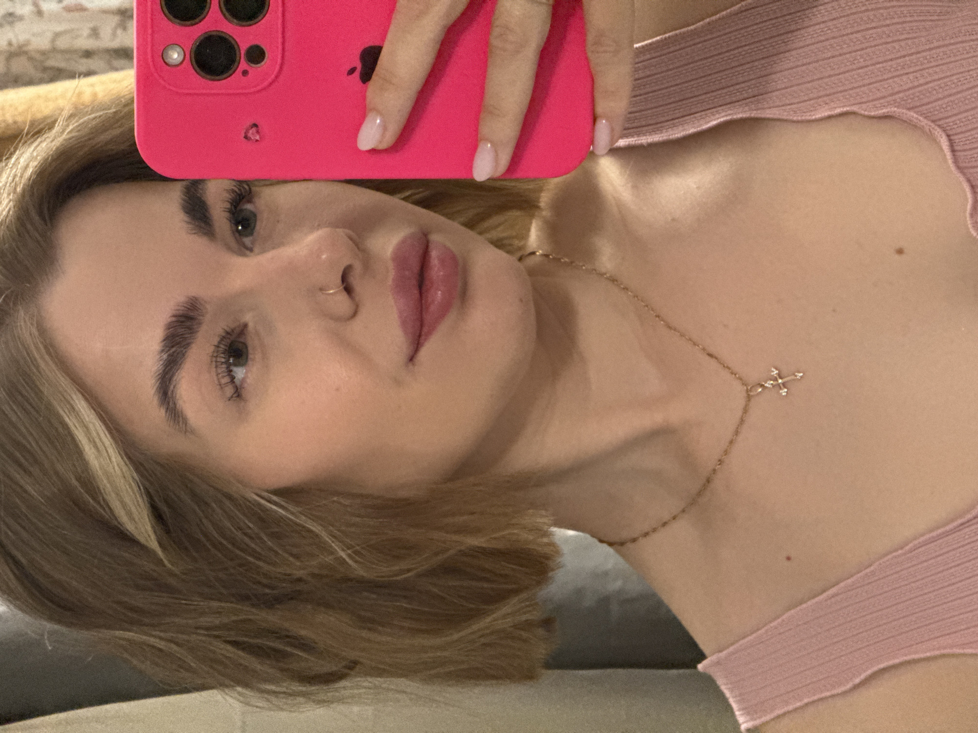 A woman with light brown hair and fair skin is holding a pink mobile phone up to her face in a selfie. She is wearing a light pink top and a delicate gold necklace with a cross pendant. A small gold hoop is visible in her left nostril. The background is blurred and indistinct, suggesting an indoor setting. There is no information available about the location being in Chaikovsky, Russia.