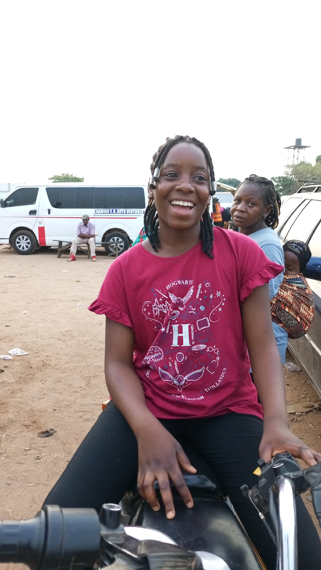 A medium shot captures a smiling young woman with braided hair and a burgundy Hogwarts t-shirt seated on a motorcycle. Her dark leggings are visible, and her hands rest on the motorcycle's handlebars and seat. Behind her, a woman with similar braids looks directly at the camera, standing next to a silver car where a child is bundled in a colorful fabric. To the left, a white van labeled "HAMISU V.A. AUTO VENTURES" is parked, and a man in a pink shirt sits on a wooden bench nearby. The ground appears sandy or dirt-covered. In the background, a water tower is visible. The image was taken outdoors during daylight. No location is shown.