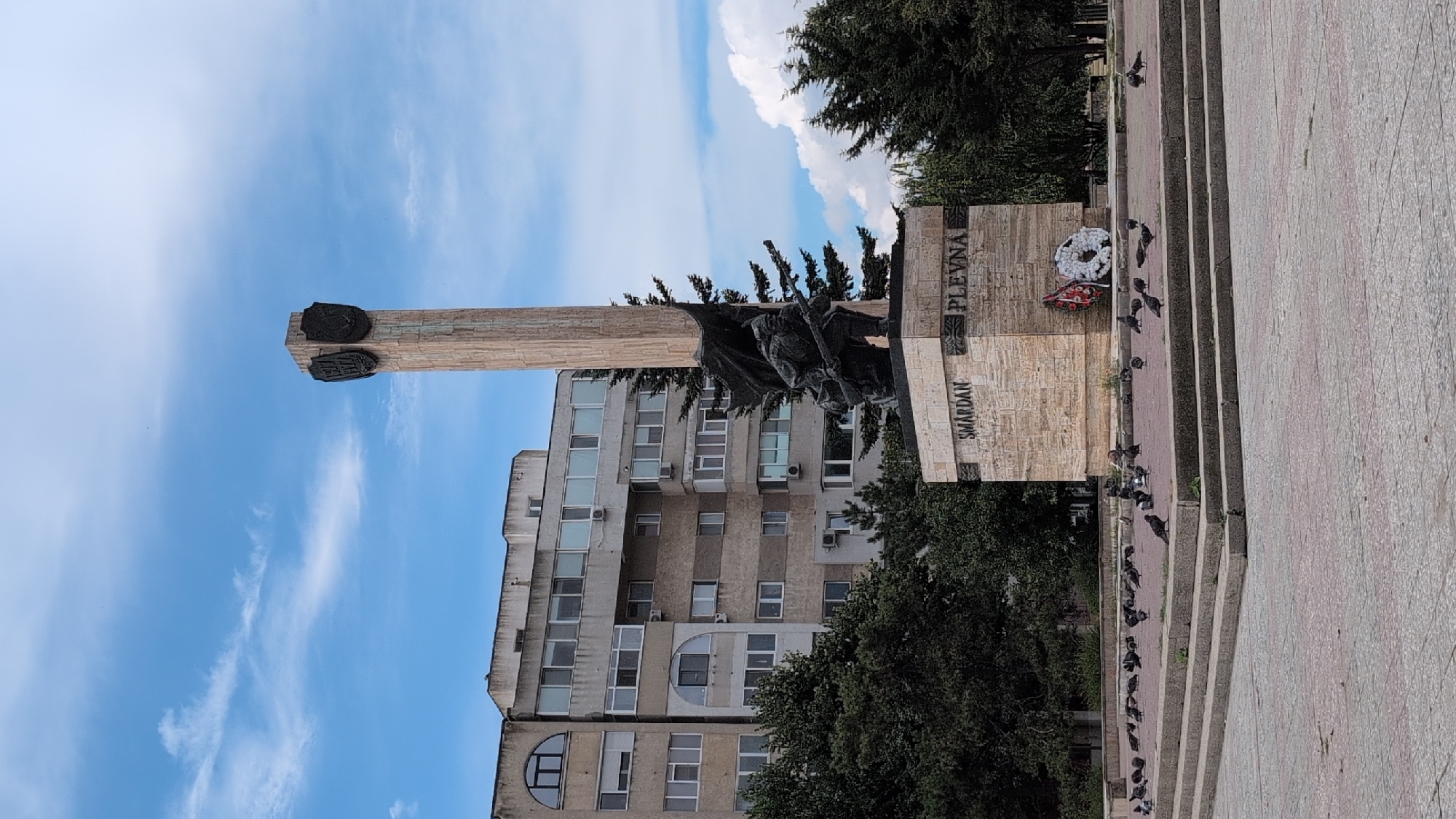 The image shows a monument with a tall, fluted column topped by two shields, and a sculpture of a soldier in a crouched position holding a rifle. The base of the monument has the words "SMÂRDAN" and "PLEVNА" carved into it. A wreath of white flowers and a red ribbon is attached to the base. The monument is situated in a public square with a checkered pavement and several steps leading up to a raised platform where the monument stands. A flock of pigeons is gathered on the platform. Behind the monument, there is a large, grey, multi-story apartment building with numerous windows. Trees line the area around the monument and in front of the building. The sky is blue with scattered white clouds. This is the Plevna Monument, located in Constanța, Romania.