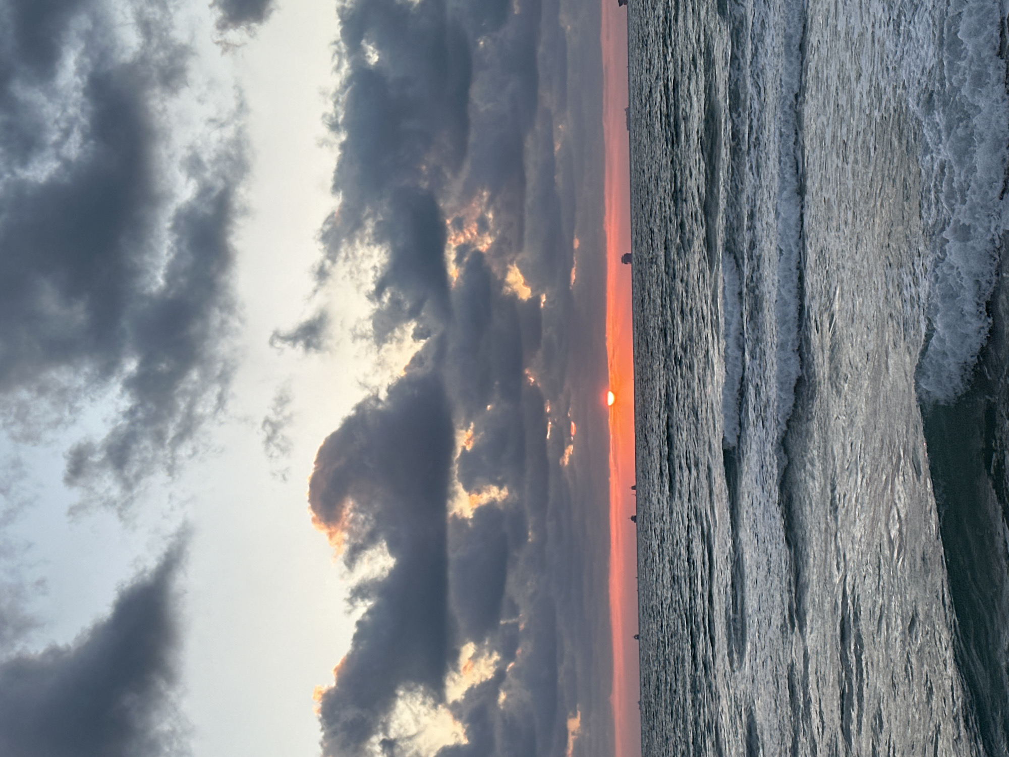 This media file depicts a seascape at sunset, oriented vertically. The image is divided into two main sections: the sky and the sea. The sky is filled with dramatic, dark clouds interspersed with patches of bright, warm light, suggesting the sun is setting behind them. The sun itself is visible as a small, orange disc peeking through a gap in the clouds near the horizon. The sea occupies the right side of the frame and displays active, choppy waves with white foam crests, indicating a dynamic water surface.
In the distance, along the horizon where the sea meets the sky, a faint silhouette of a skyline or landmass can be observed, dotted with several small, dark shapes that might be ships or structures. The overall mood conveyed is one of natural beauty and perhaps a touch of drama due to the stormy-looking clouds and the vibrant sunset colors. The time of day is clearly sunset, and the weather appears to be mixed, with dark clouds but also revealing sunshine.
The location context suggests this scene is likely near Haifa, Israel, a coastal city. While there is no visible text in the image, the elements present - the sea, the sunset, and the distant horizon with possible maritime elements - combine to create a serene yet powerful natural vista.