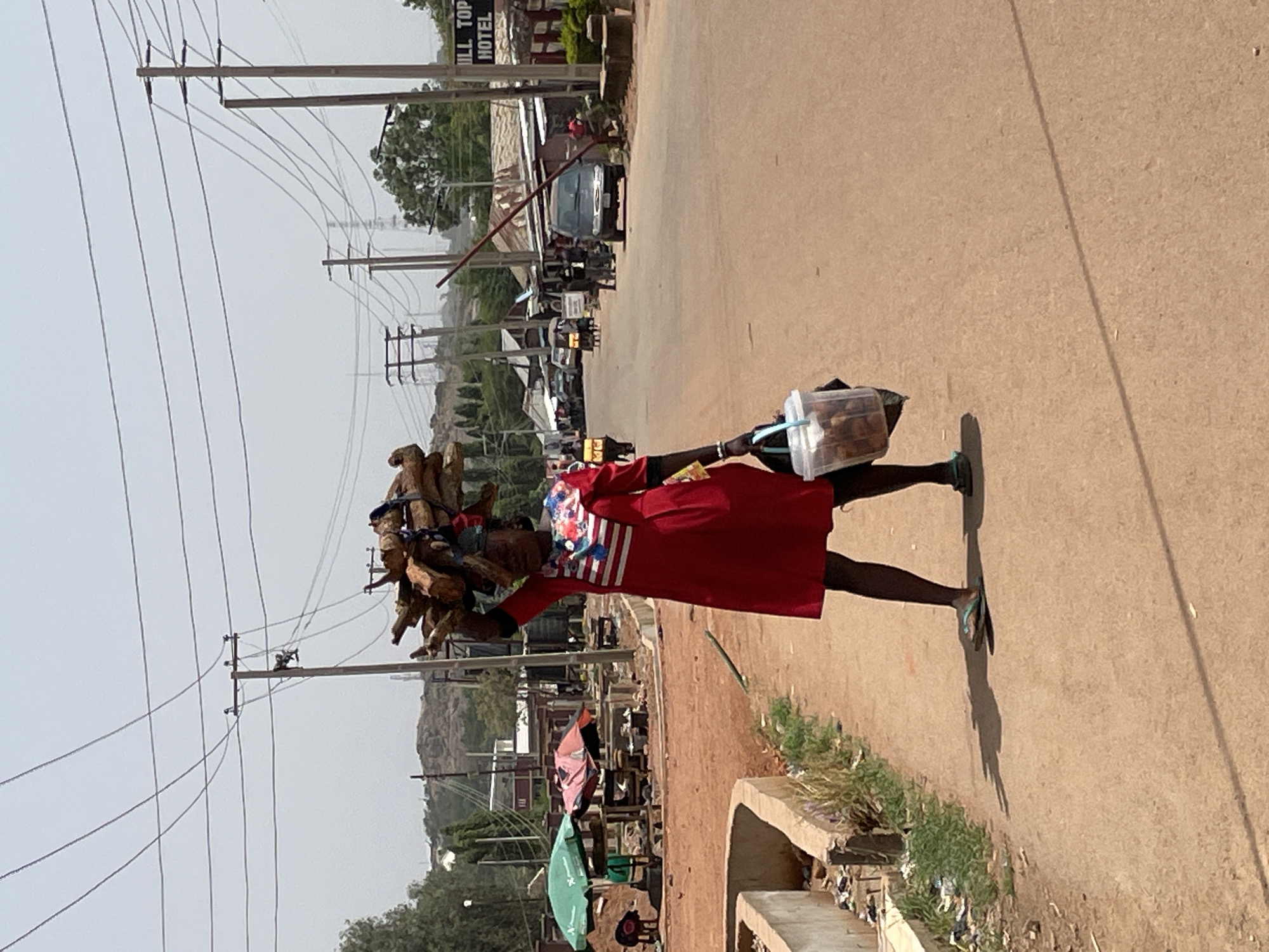 A man walks along a road in Jos, Nigeria. He balances firewood on his head and carries a container filled with items in his right hand. He is wearing a red dress with white stripes around the neck. The road he walks on is lined with roadside businesses and utility poles. A sign for "HILL TOP HOTEL" is visible in the background.