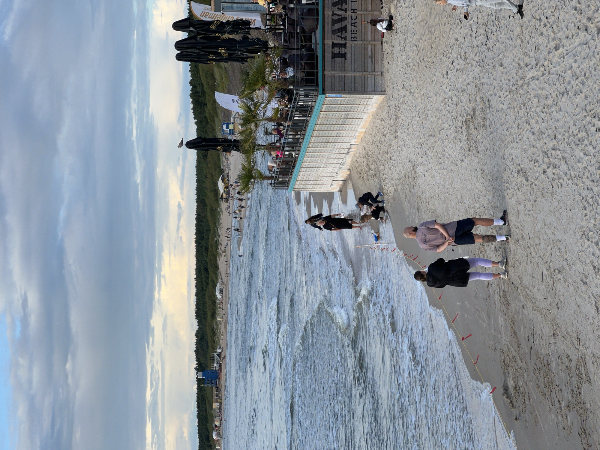 A wide-angle, eye-level shot captures a bustling beach scene on a cloudy day, likely in Palanga, Lithuania, given the presence of the "Havana Beach Bar" visible in the image. In the foreground, a sandy beach stretches out, marked by a yellow rope with small red flags. Two individuals stand facing the ocean, a woman in a black jacket and light purple leggings, and a man in a grey t-shirt and dark shorts. To the right of the frame, a wooden building with a turquoise trim houses the "Havana Beach Bar," its outdoor seating area filled with patrons enjoying the atmosphere. Palm trees are interspersed with black umbrellas, adding a tropical touch. Further into the background, more beachgoers stroll along the shoreline, and a distinctive blue lifeguard tower stands prominently near the treeline. The ocean, with its gentle waves, extends towards the horizon, where the cloudy sky meets the distant treeline. Several people are scattered across the sand, some seated, others walking or playing. A seagull perches atop a closed black umbrella, surveying the scene. The overall impression is one of a lively recreational area by the sea, with a mix of relaxation and activity.