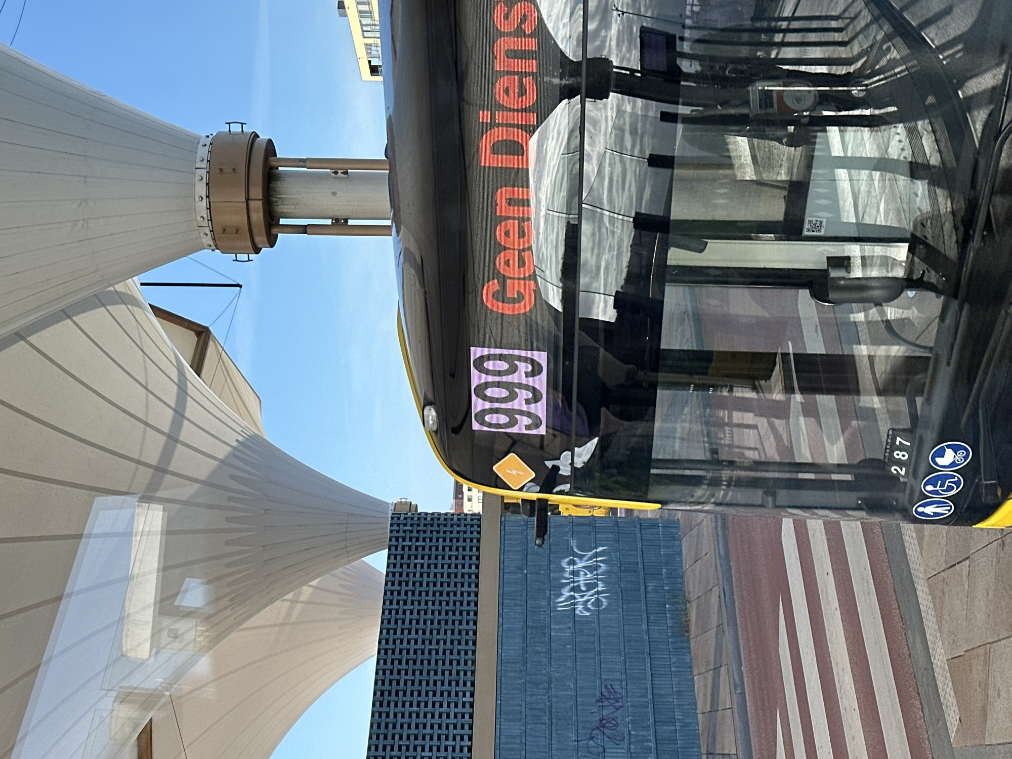 The image shows the front of a yellow and black bus at a bus stop. The bus has a digital display showing "999 Geen Dienst", meaning "999 No Service". The bus stop has a modern, covered structure with white, sail-like canopies. Behind the bus, a blue brick wall with some graffiti is visible. To the left of the bus, a crosswalk with red and white stripes is present. The sky is blue and clear, suggesting it is daytime. The bus also displays symbols indicating accessibility for people with disabilities and for strollers. The number "287" is visible on the bus, likely its route or vehicle number. The scene appears to be a public transportation hub in Utrecht, Netherlands.