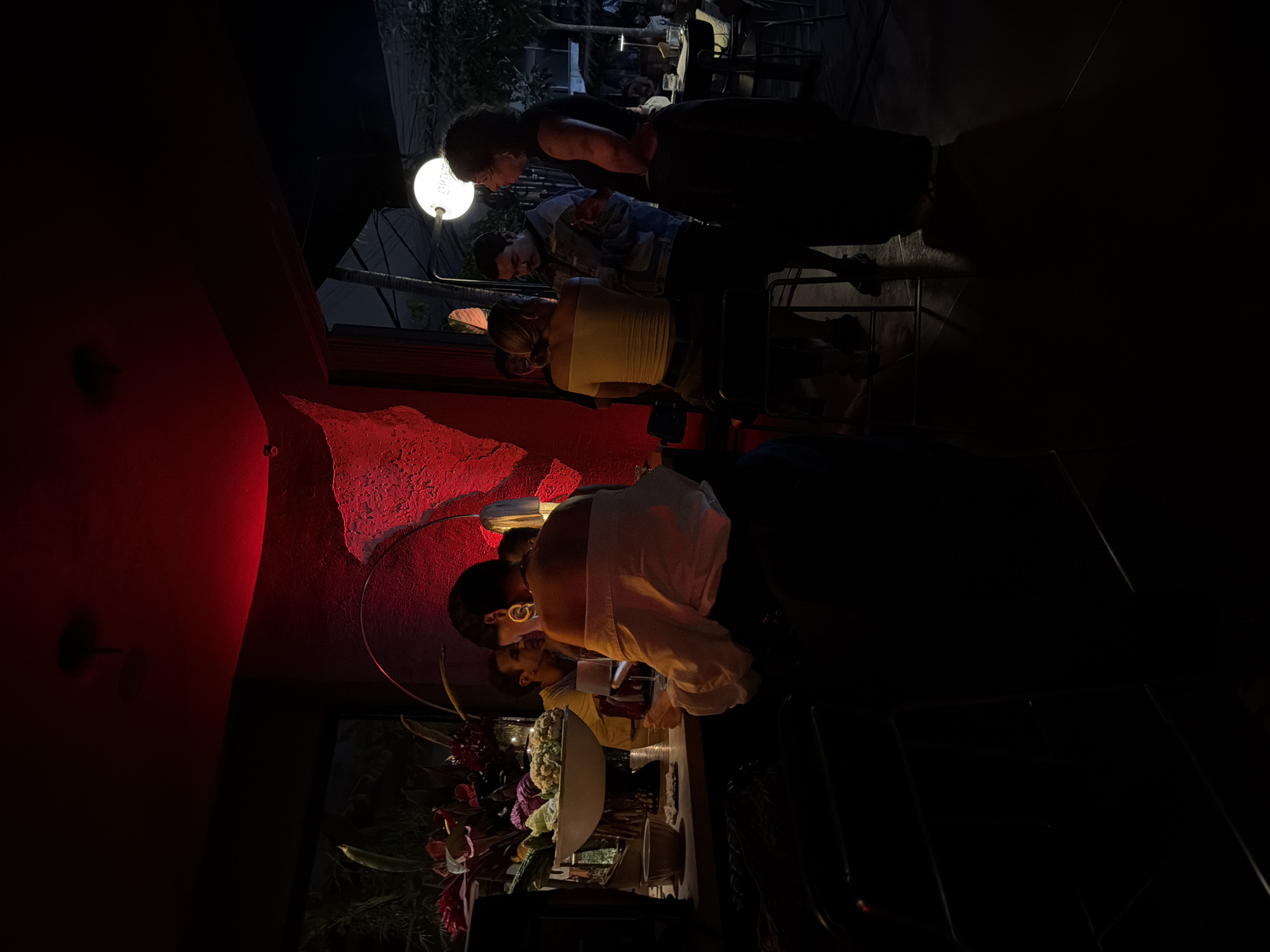 In a dimly lit establishment in Mengwi, Indonesia, a group of individuals are gathered. The space is characterized by a prominent red-lit textured wall, suggesting a modern or artistic interior design. On the left, a bar area is visible with floral arrangements and a bowl of vegetables. Several people are interacting at or near the bar. In the foreground, a woman with short dark hair, wearing an off-the-shoulder white top and dark pants, is seated with her back to the camera, facing a table. To her right, another woman with a yellow top and dark pants is seated on a stool. Further back, a man in a denim jacket is speaking with a woman wearing a black top and dark skirt, who is facing the camera. Another man is partially visible behind the woman in the yellow top. In the background, a bright circular light fixture hangs, and through an open doorway, an outdoor scene with trees and more lighting is discernible. The atmosphere appears to be social, possibly a bar or restaurant setting.