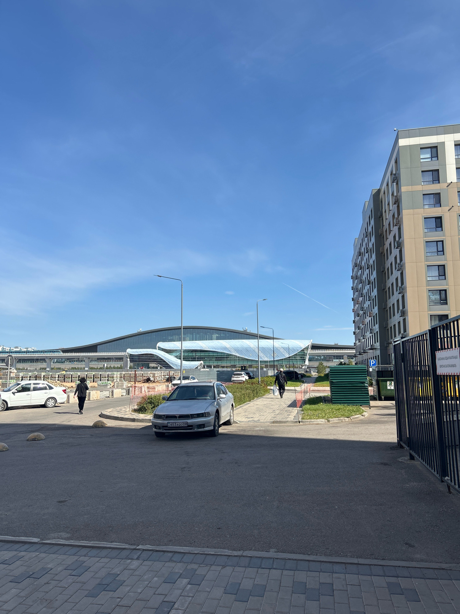 An eye-level outdoor shot shows a paved area in front of a modern apartment building and a large airport terminal. The sky is clear blue with a faint contrail. In the foreground, a silver sedan is parked. Further back, a white sedan is driving away. A person in a black jacket and pants is walking on a sidewalk towards the apartment building. The apartment building has a beige and grey facade with multiple windows. The airport terminal in the background features a distinctive curved glass roof. A black metal fence is visible on the right side of the image, with a sign in Russian attached to it. The scene appears to be in Astana, Kazakhstan, likely near Nazarbayev International Airport.