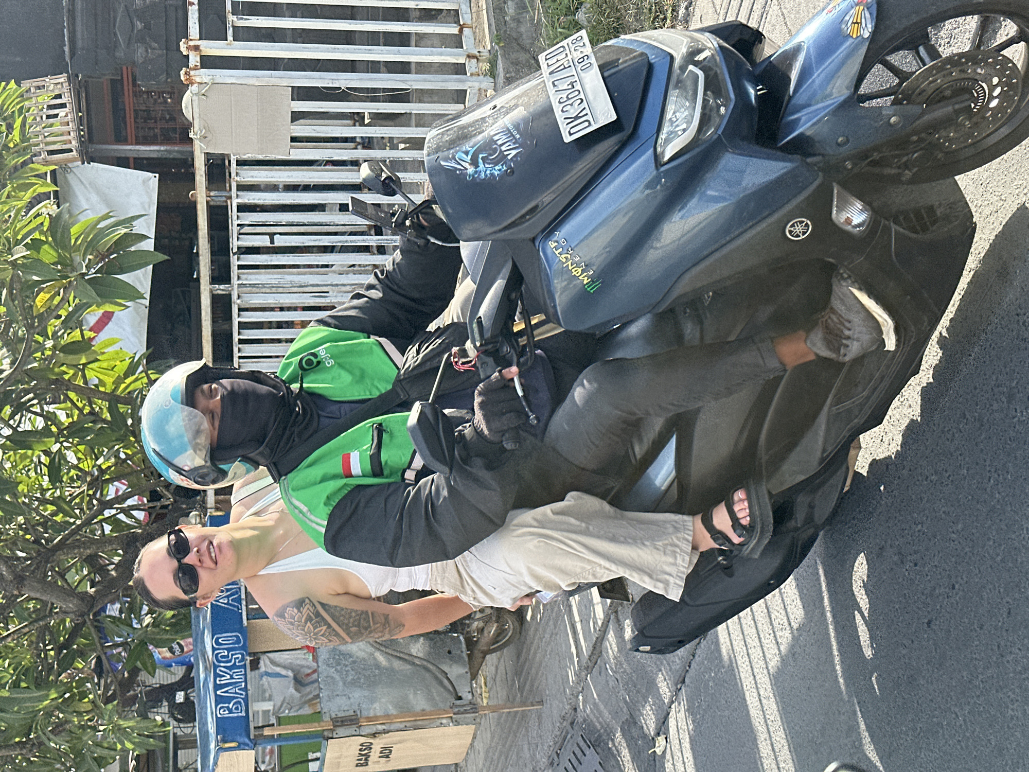 In North Kuta, Indonesia, a woman with sunglasses and a white tank top is riding as a passenger on a blue Yamaha scooter. She has a large tattoo on her left arm. The driver wears a green and black jacket with a red and white stripe, a black face mask, and a light blue helmet. A food cart with the text "BAKSO ADI" is visible in the background. The scooter has a license plate that reads "DK 3647 AED" and a "Monster Energy" sticker on the front. A white metal fence is also present.