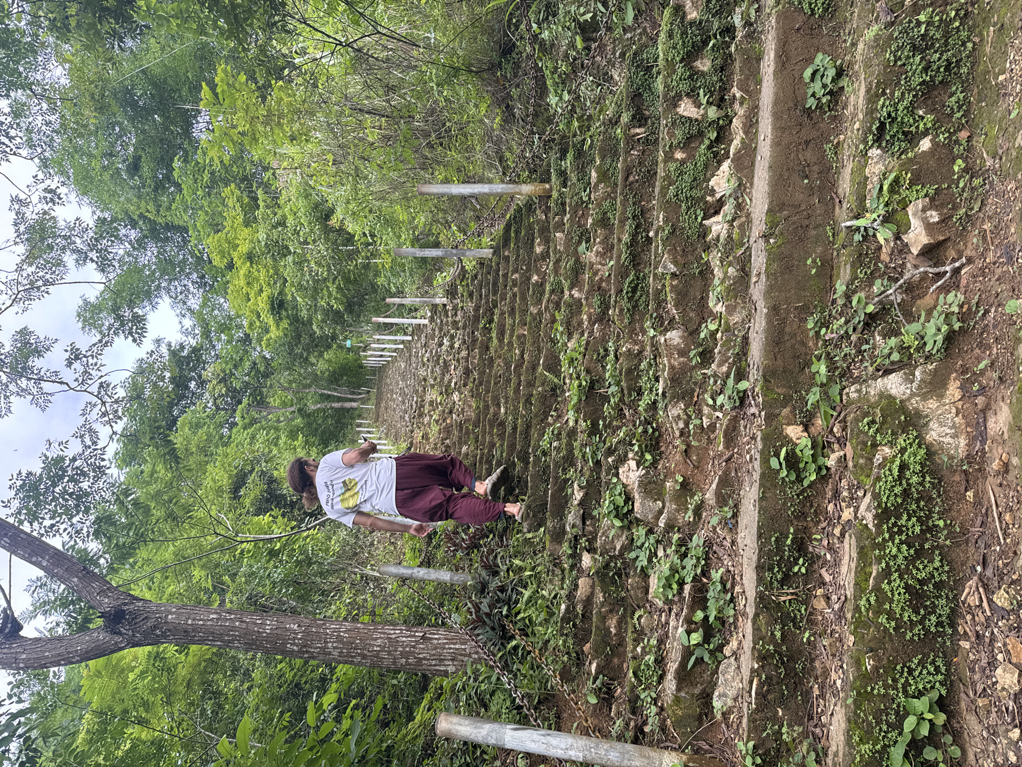 A person walks up a stone staircase in a lush green environment, possibly Xóm Khán, Vietnam. The staircase is lined with metal posts and chains on the left side, and the steps are uneven and covered with moss and vegetation. The person is wearing a white t-shirt and loose-fitting maroon pants, with their back to the camera. They are barefoot, wearing sandals. The surrounding area is dense with trees and foliage, suggesting a natural or semi-natural setting. The sky is overcast.