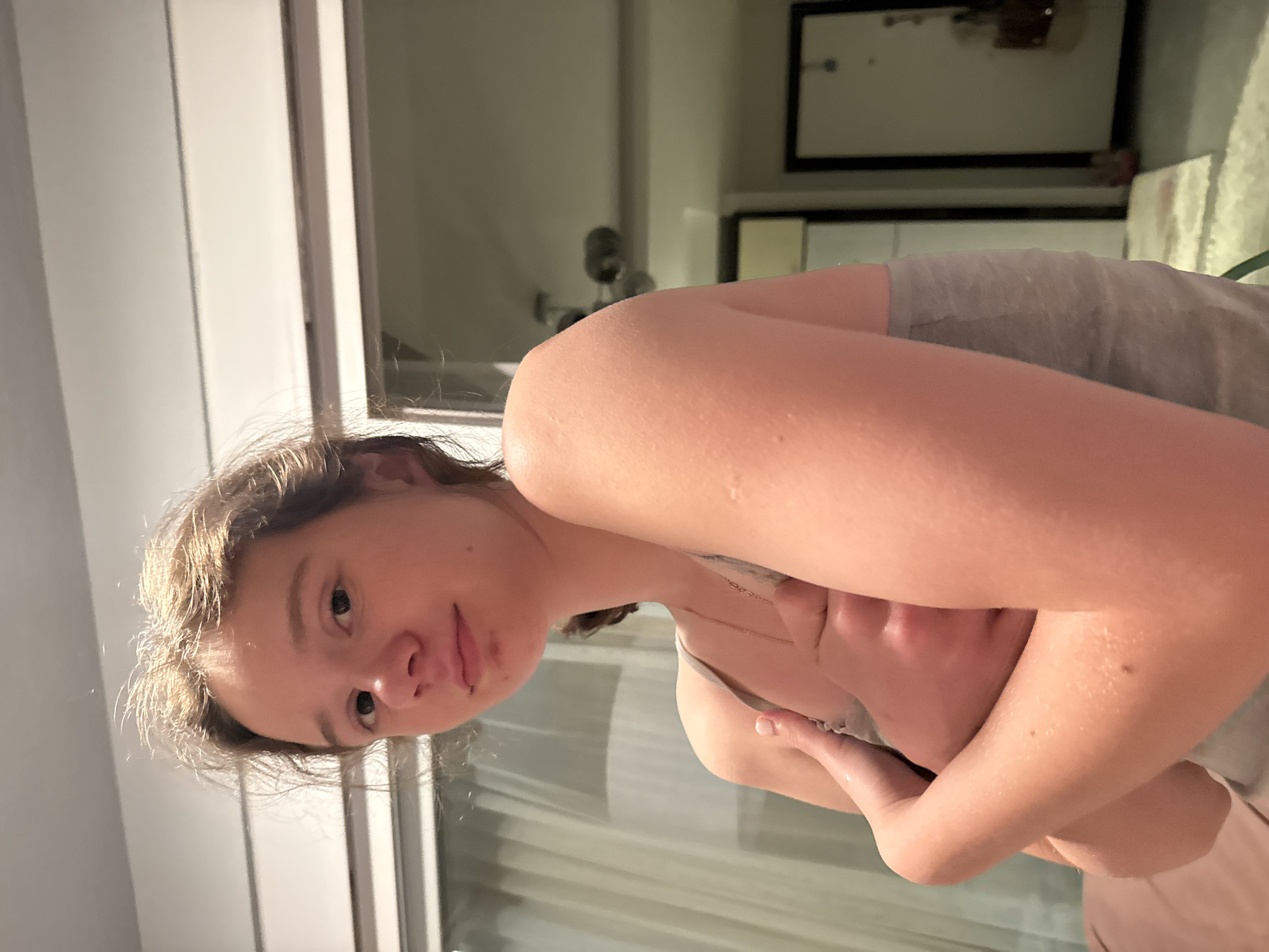 A young woman is holding her arms up in front of her face, looking at the camera. She is wearing a grey tank top. Behind her is a window with white blinds. In the background, there is a framed mirror. The location is Unknown, Unknown.