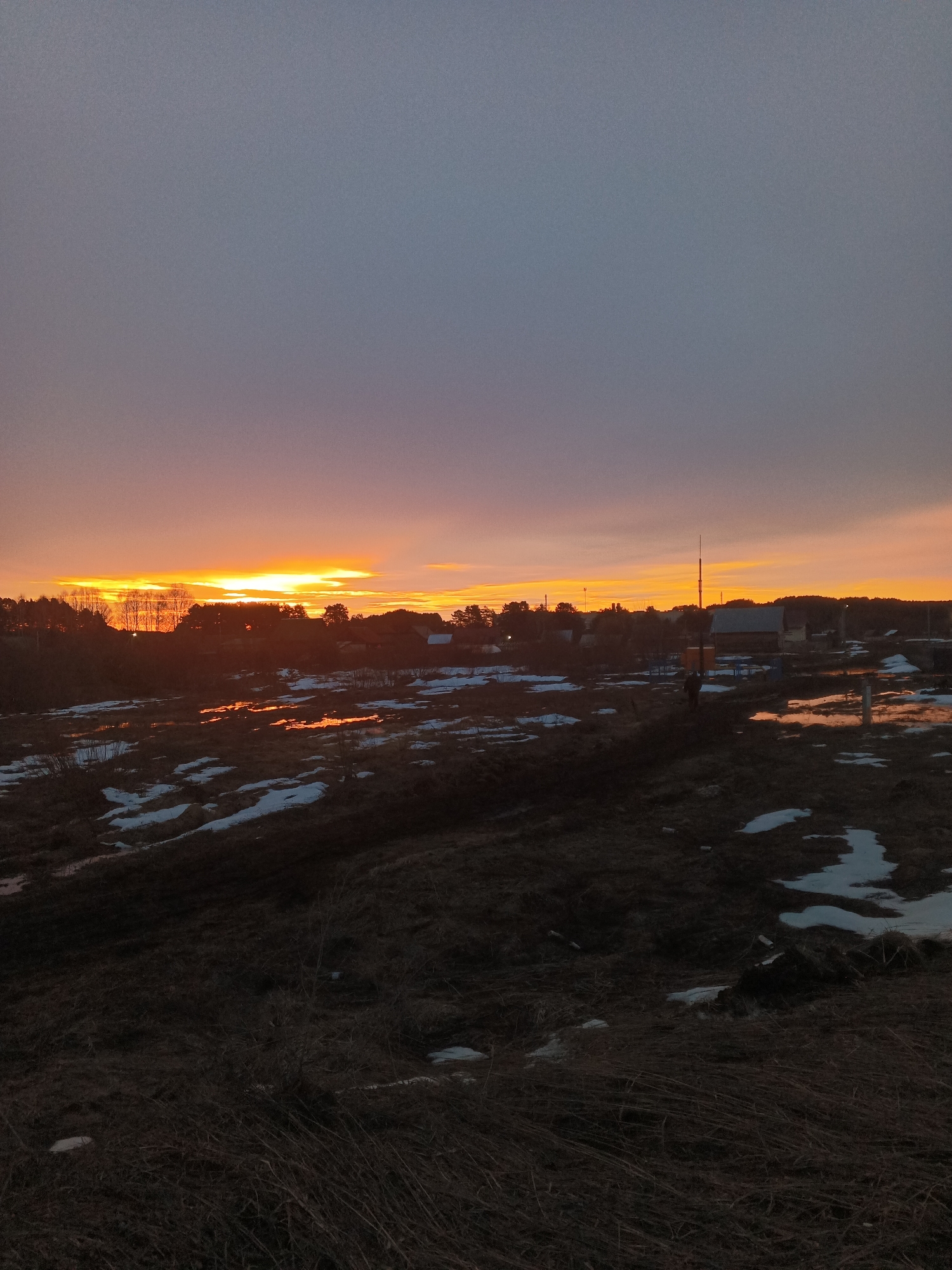 The image depicts a landscape at sunset in what appears to be the rural area of Rusalevka, Russia. The sky is filled with hues of orange and purple, typical of a sunset. The horizon is silhouetted with trees and possibly buildings. The foreground features a field with patches of melting snow and pools of water reflecting the colors of the sky. The ground is predominantly brown, with dry grass visible. No people are clearly visible in the image.