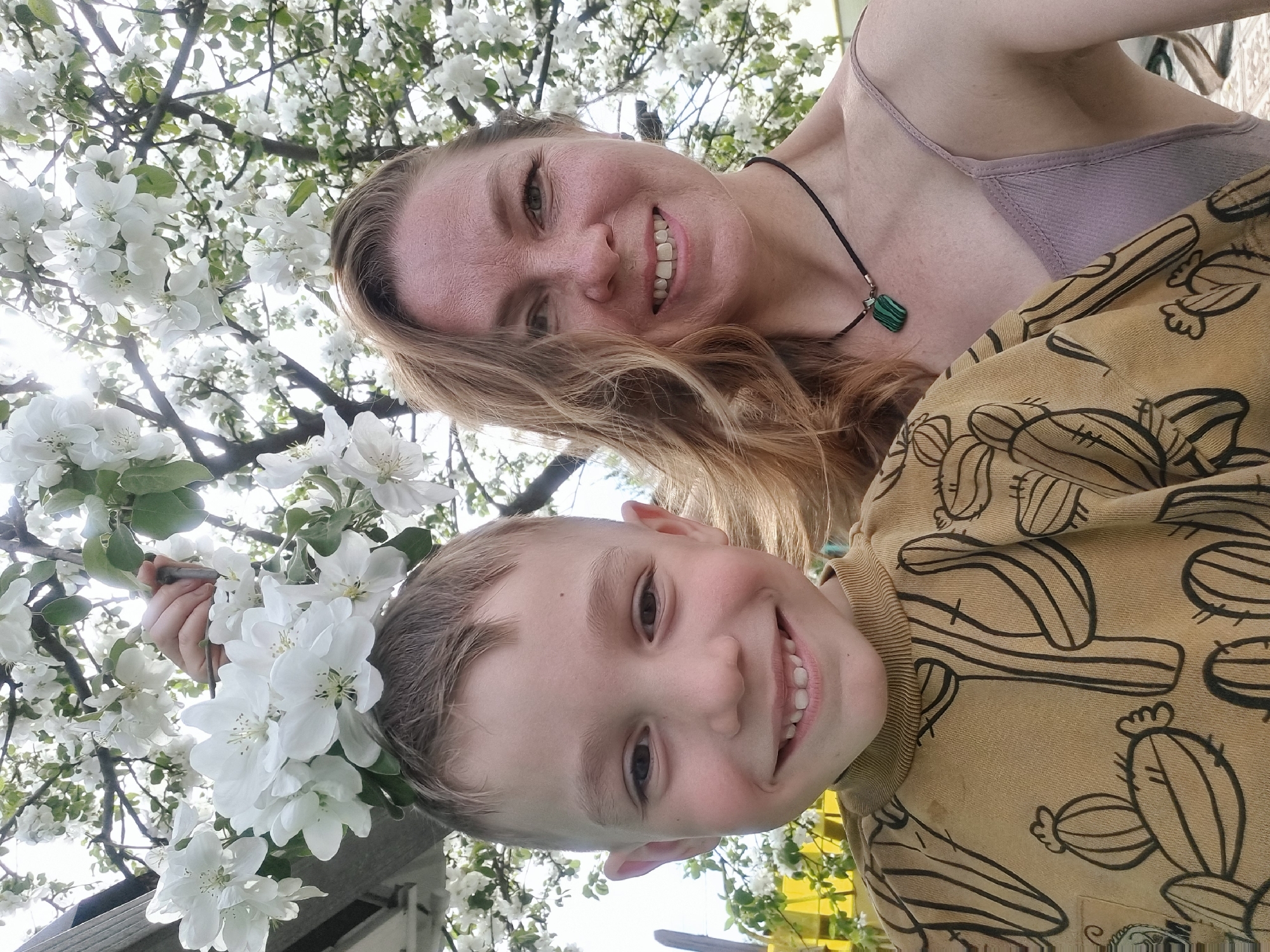 A photo captures a woman and a young boy smiling while standing under a flowering tree. The woman wears a light-colored top and a black necklace with a green pendant. The boy is wearing a yellow shirt with black cactus print. They are both smiling, and white flowers from the tree surround them. The photo was taken outdoors, and the background suggests a garden or park-like setting, possibly in Chaikovsky, Russia, although there are no explicit indicators pointing to that specific location in the image.