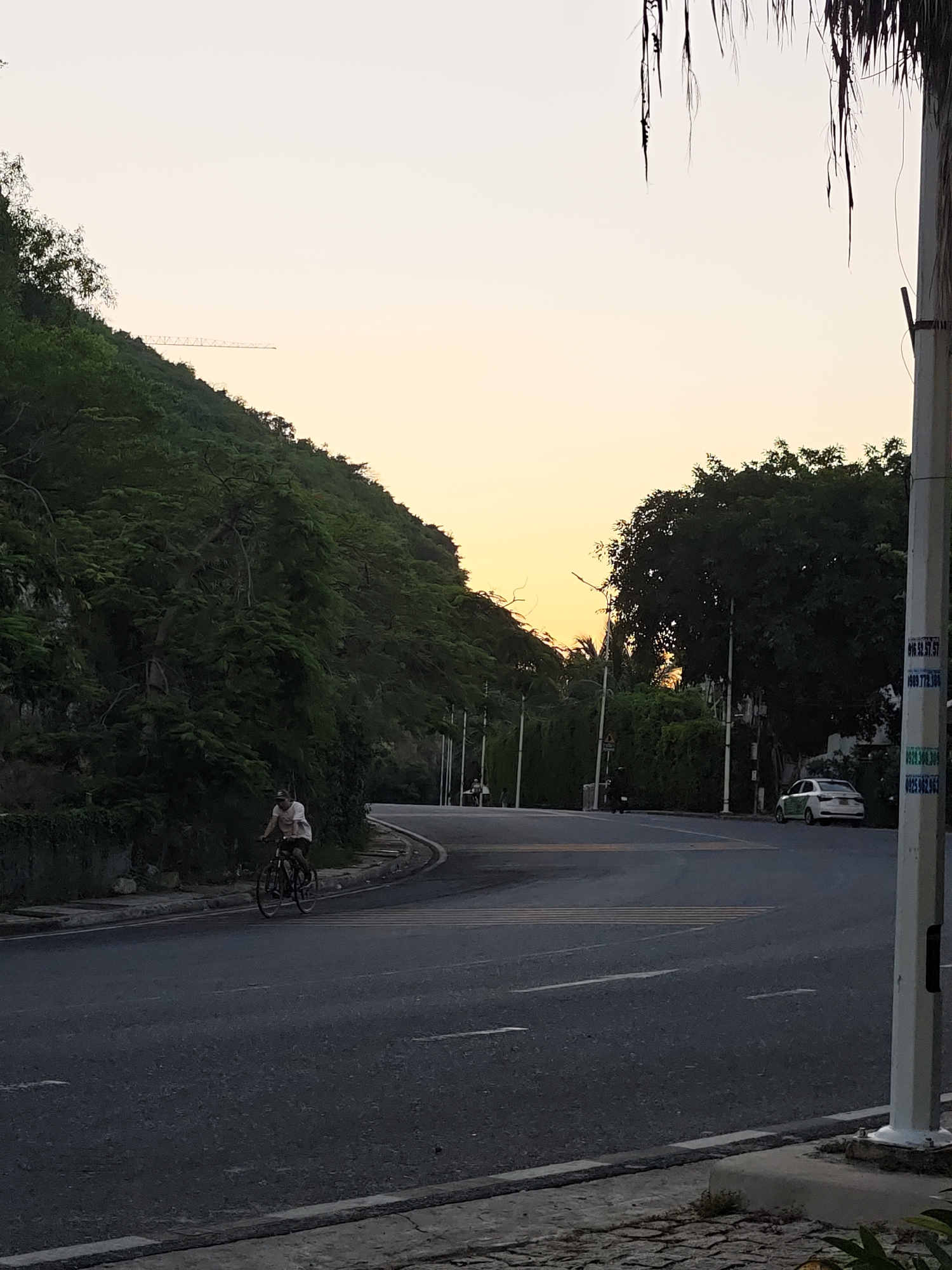 A person rides a bicycle on a paved road in Bac Nha Trang Ward, Vietnam, as dusk settles. The road curves to the right, with a steep, tree-covered hill on the left and a line of trees and buildings on the right. The sky is a pale orange and yellow, indicating the setting sun. A white car with green accents is parked on the right side of the road. A lamppost with signage is visible in the foreground on the right. The bicycle rider is wearing a light-colored t-shirt and a hat, and the bicycle is dark-colored. The overall scene depicts a tranquil evening commute or recreational ride in a coastal city.
