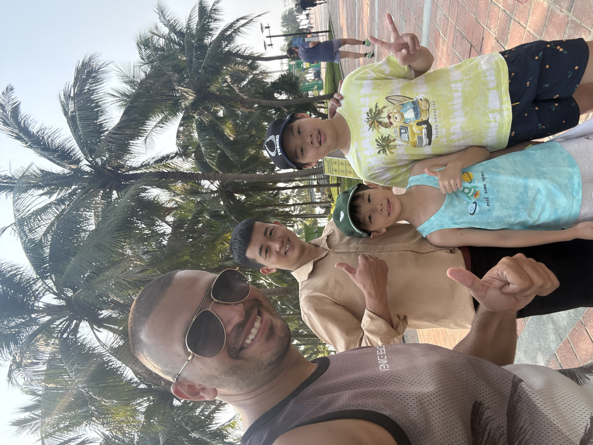 The image shows a selfie taken outdoors, likely in a coastal area in Đà Nẵng, Vietnam. Four individuals are visible: a man with sunglasses in the foreground, presumably taking the picture; an adult male behind him; and two younger boys above them. The boys are making hand gestures, one with a peace sign, the other with a thumbs-up. All are smiling. The background features palm trees and a paved walkway. A person is jogging in the distance.