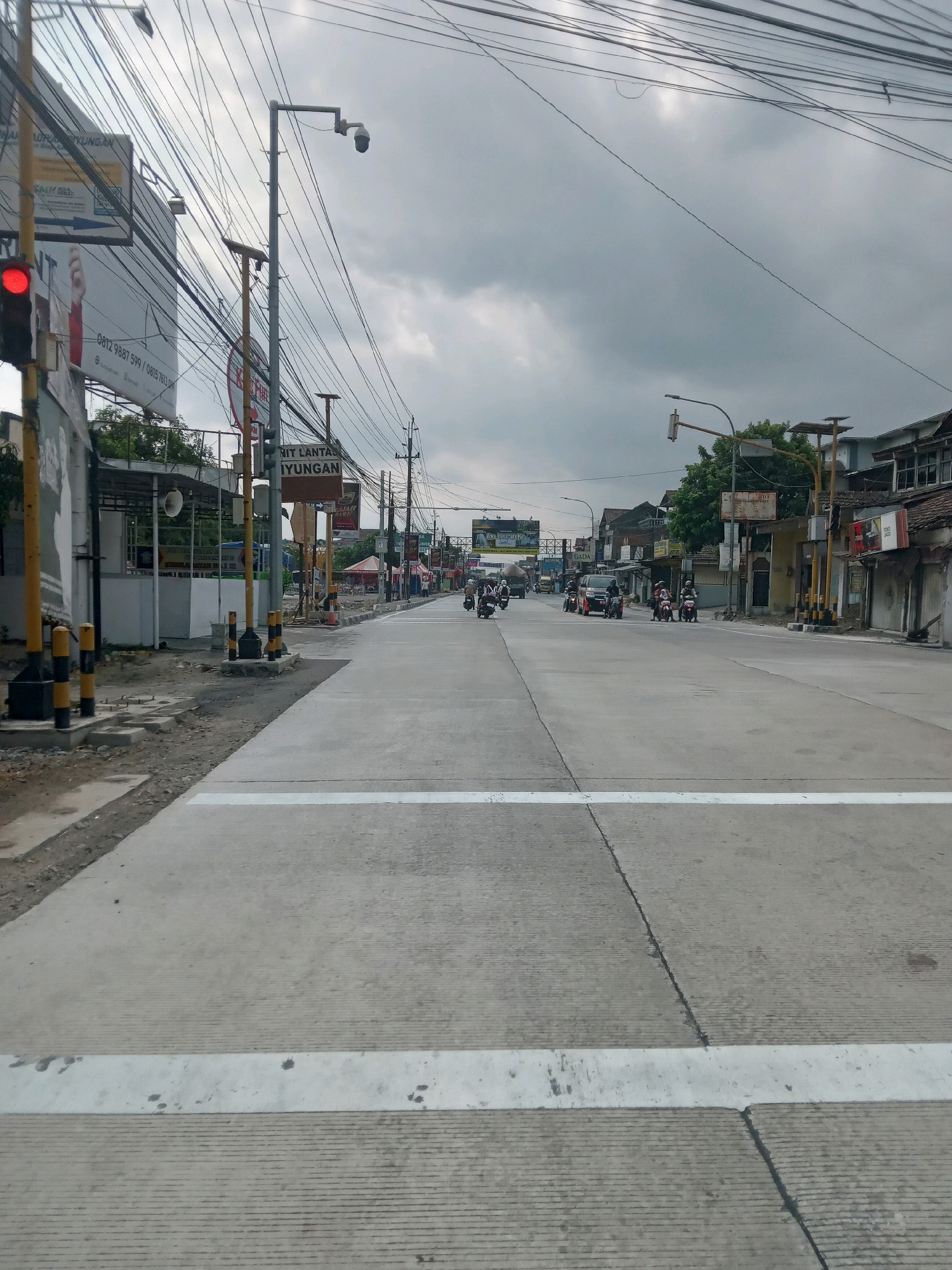 The image shows a street scene in Srimartani, Indonesia, under an overcast sky. The road is paved with concrete and has white pedestrian crossing lines. Several motorcycles and a few cars are visible on the road, with some vehicles stopped at what appears to be an intersection controlled by a traffic light, which is currently red. Utility poles with numerous overhead wires are a prominent feature along the sides of the street, along with streetlights and surveillance cameras. Buildings line the street, some appearing to be commercial establishments with signage. Trees are present in some areas. The overall impression is of a typical urban or semi-urban street in Indonesia with ongoing traffic.